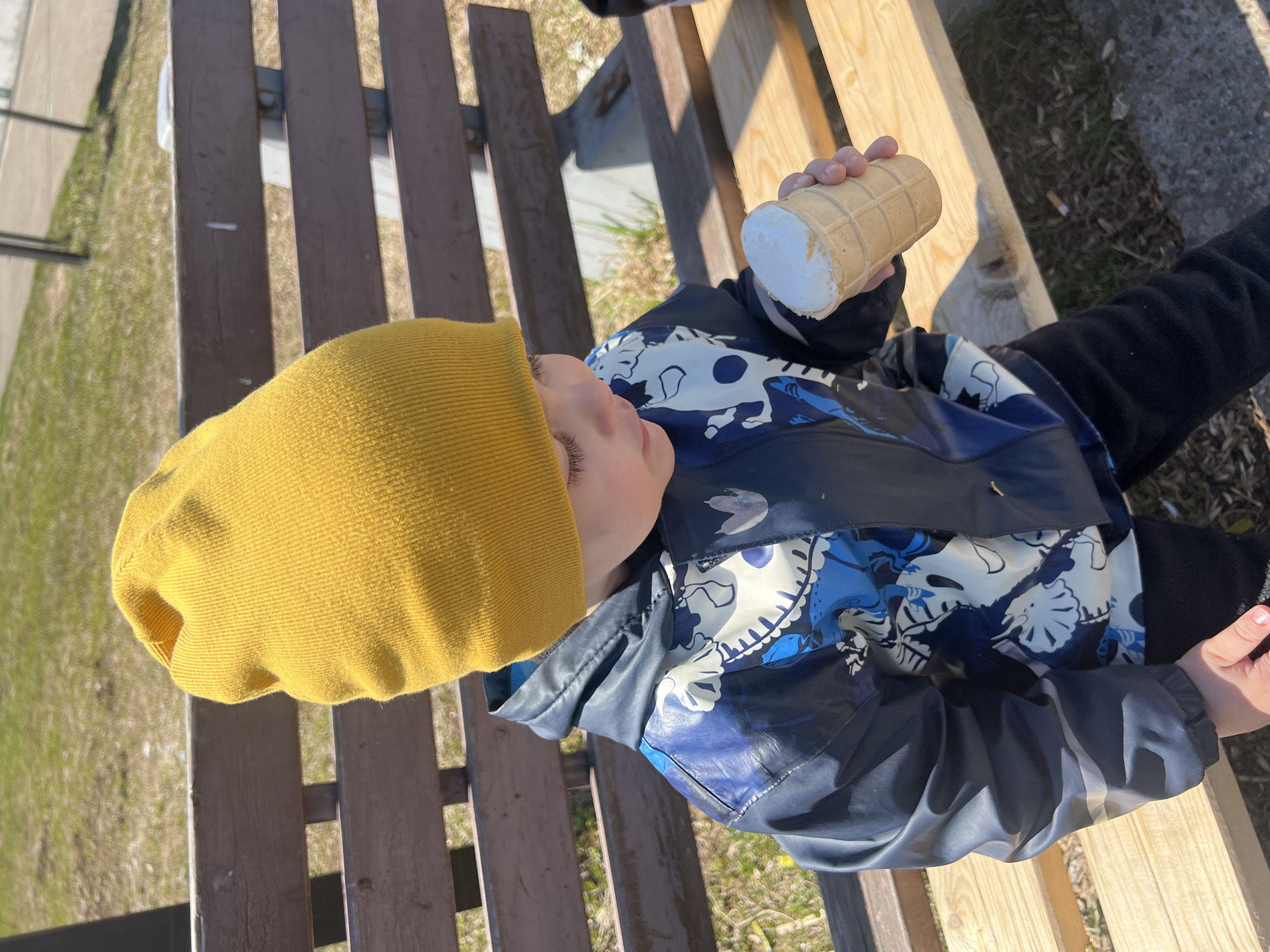 A young child, wearing a yellow beanie and a blue and white patterned jacket, sits on a wooden bench outdoors, holding an ice cream cone. The bench has brown slats. The location appears to be outdoors in an urban setting, possibly Марковский, Russia, based on the user's location. The child's gaze is directed upwards.