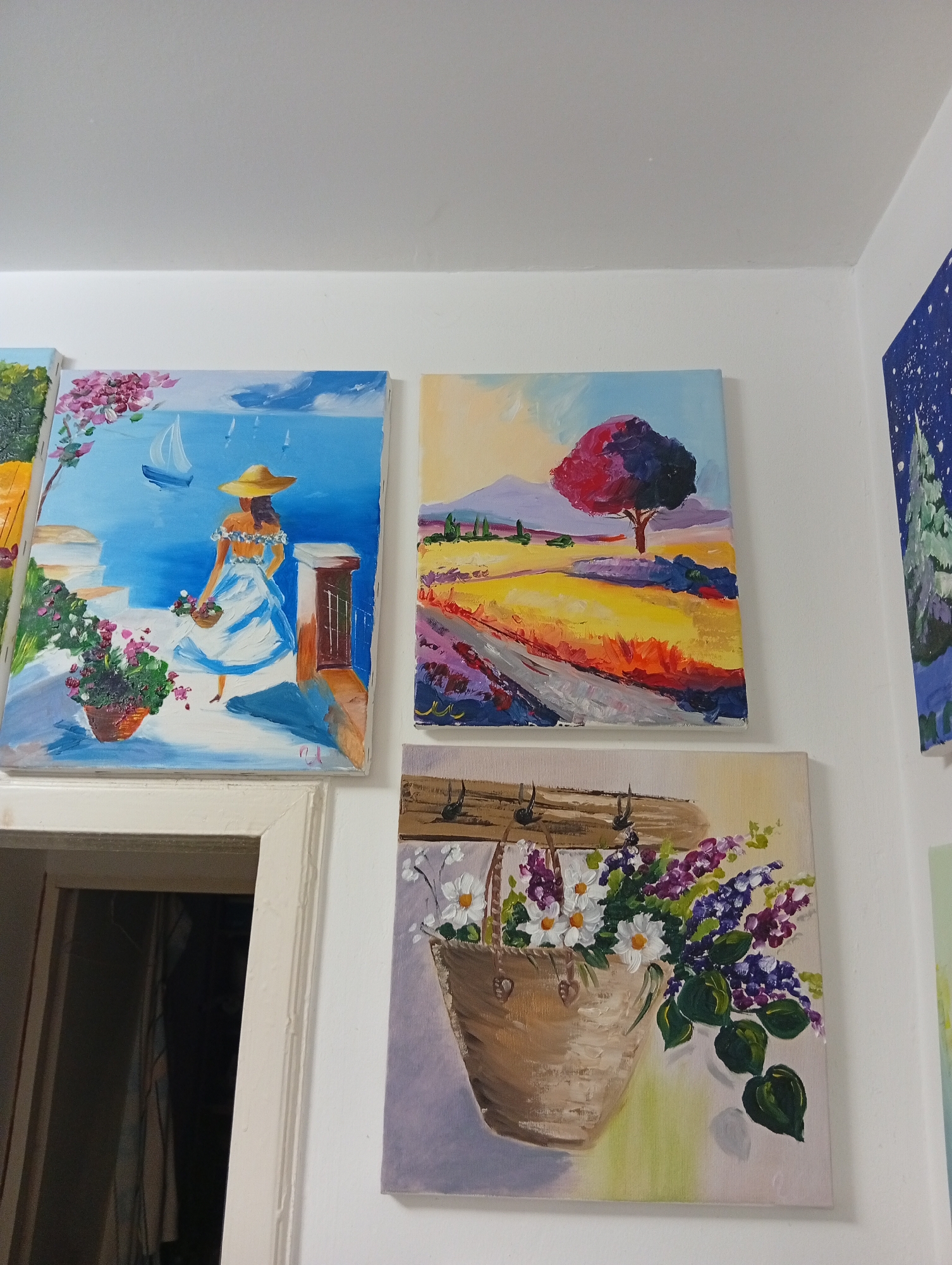 A medium, eye-level shot shows a section of a white wall decorated with four paintings. To the left of the frame, a partially visible doorway leads into a dark room.
The first painting, positioned in the top left, depicts a woman in a white and blue dress and a wide-brimmed straw hat standing on a terrace overlooking the sea. A sailboat is visible in the distance. A pot of pink flowers adorns the terrace.
The second painting, to the right, shows a landscape with a red-leafed tree, a field of yellow and purple flowers, and distant mountains.
The third painting, located below the second, presents a basket filled with white daisies and purple lilacs.
A portion of a fourth painting, a blue and green landscape, is visible on the right edge of the frame.
The scene appears to be indoors, but there is no information about the specific location in Varna, Bulgaria.