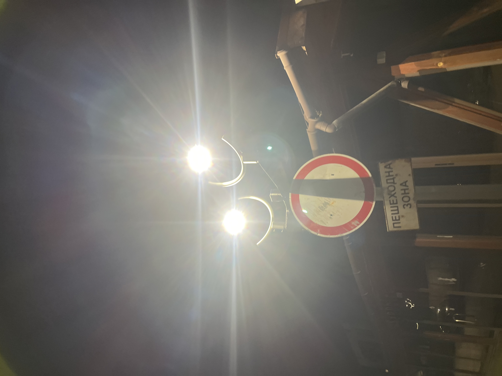 The image shows a nighttime street scene in Goce Delchev, Bulgaria. A bright streetlight illuminates a "No Entry" traffic sign, with a partially visible pedestrian zone sign to the right. The signs are attached to a pole, and there are building elements, including a drainpipe and wooden structures, in the background. The overall lighting is poor, with the streetlight causing glare.