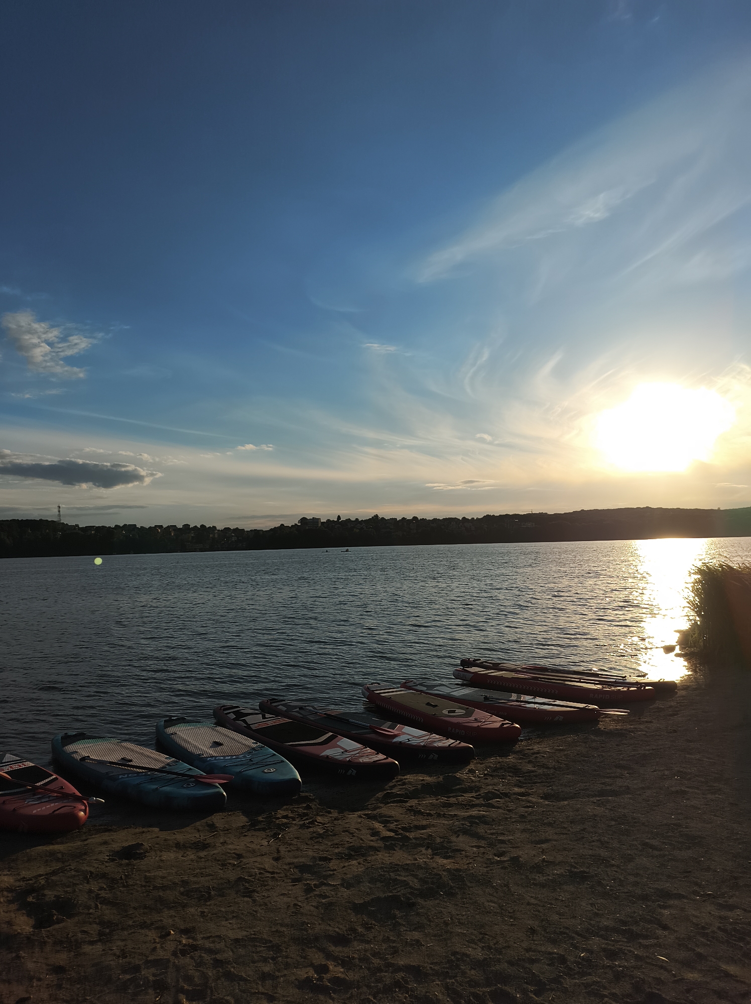 A scenic view of a lake at sunset in Ternopil, Ukraine. The sun, a bright orb low in the sky, casts a golden reflection on the water. Several paddleboards are lined up on the sandy shore in the foreground, ready for use. The distant shoreline is lined with trees and buildings. The sky is a mix of deep blue and wispy clouds, with the sun creating a brilliant glow.