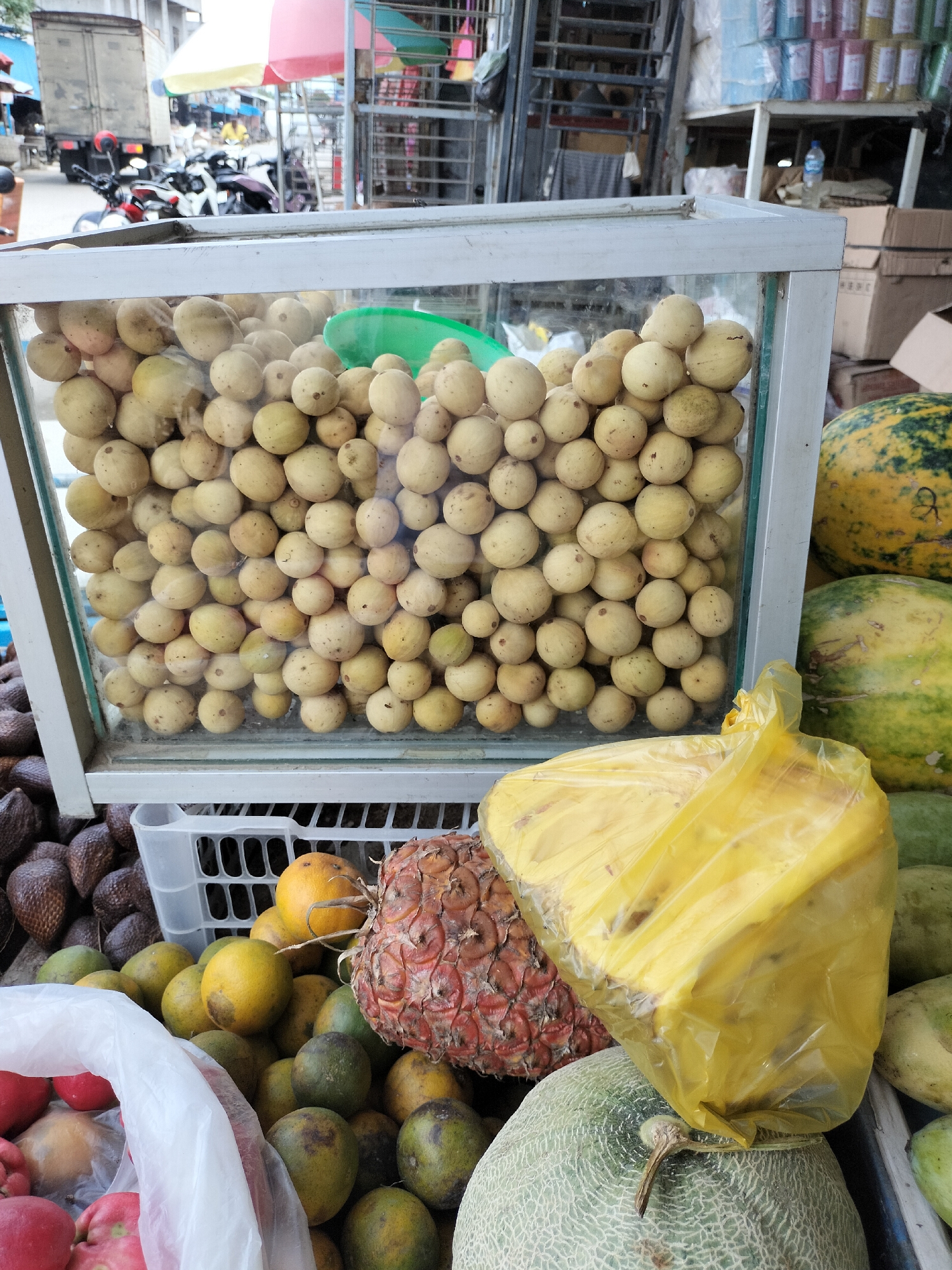 The image captures a vibrant outdoor market scene in Kotapantonlabu, Indonesia, focusing on a stall overflowing with fresh produce. In the foreground, a glass display case is filled with numerous small, pale yellow fruits, possibly langsat or duku. To the right of this display, a large, yellow plastic bag appears to contain more fruit, its contents partially obscured. Beneath the display case, a white plastic crate holds a variety of fruits, including oranges, and what seems to be a pineapple with its characteristic spiky skin. Further to the right, two large watermelons, one with distinct yellow and green stripes and the other predominantly green, are visible. In the background, stacks of goods and a parked truck suggest the bustling nature of the market. The overall impression is one of abundant, fresh food offered for sale in a lively local setting.