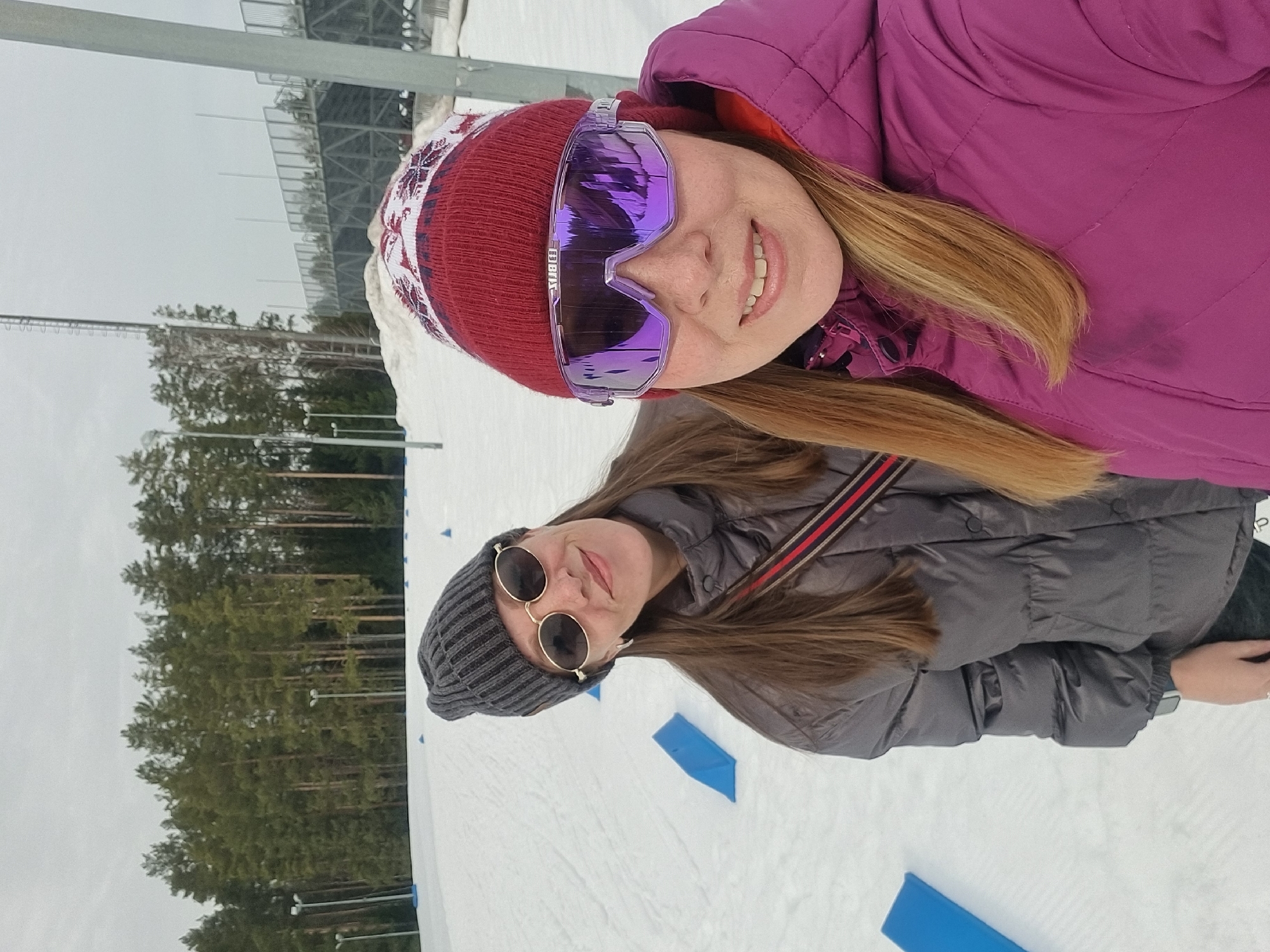 Two adult females are depicted outdoors in a snowy environment in Прикамский, Russia. The individual in the immediate foreground, positioned to the right, has light brown hair, wears a red knit beanie with white snowflake patterns, purple mirrored ski goggles, and a quilted magenta winter jacket, smiling towards the camera. The second individual, positioned slightly behind and to the left of the first, has long dark brown hair, wears a grey knit beanie, round dark-lensed sunglasses with gold frames, and a grey quilted winter jacket. A black bag with a red and white striped strap is visible across her chest. She is also smiling. The ground is covered in white snow, with several small, blue triangular markers arranged across the surface. In the background, a dense line of evergreen trees is visible. Beyond the trees and to the far right, tall light poles and a multi-level metal structure resembling stadium seating or a viewing platform are present. The sky is overcast and light grey.