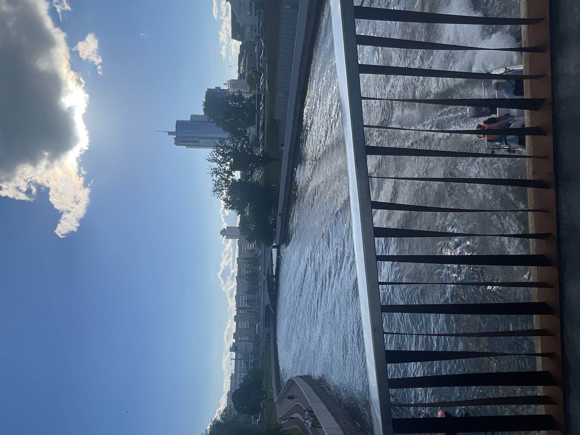 This is a vertical, slightly angled, eye-level shot of a river running through a city on a sunny day. The foreground is dominated by a dark metal railing with wooden accents, suggesting a bridge or elevated walkway. Through the railing, the river is visible, with small ripples and a boat in the distance creating white spray. The boat appears to have at least two people on it, one wearing an orange life vest.
Beyond the river, the scene opens up to a cityscape. Tall, modern buildings with glass facades rise into the sky, interspersed with trees. The sky is a vibrant blue, with a large, bright cloud on the left that partially obscures the sun, creating a lens flare effect. The sun's rays are strong, casting reflections on the water and illuminating the clouds with a slight iridescence. Along the riverbank, a paved walkway is visible with people strolling.
The overall impression is one of a lively urban environment enjoying a pleasant, sunny day. The time of day appears to be late morning or early afternoon, given the brightness of the sun and the clear blue sky. The weather is fair and warm. No discernible text is visible in the image.