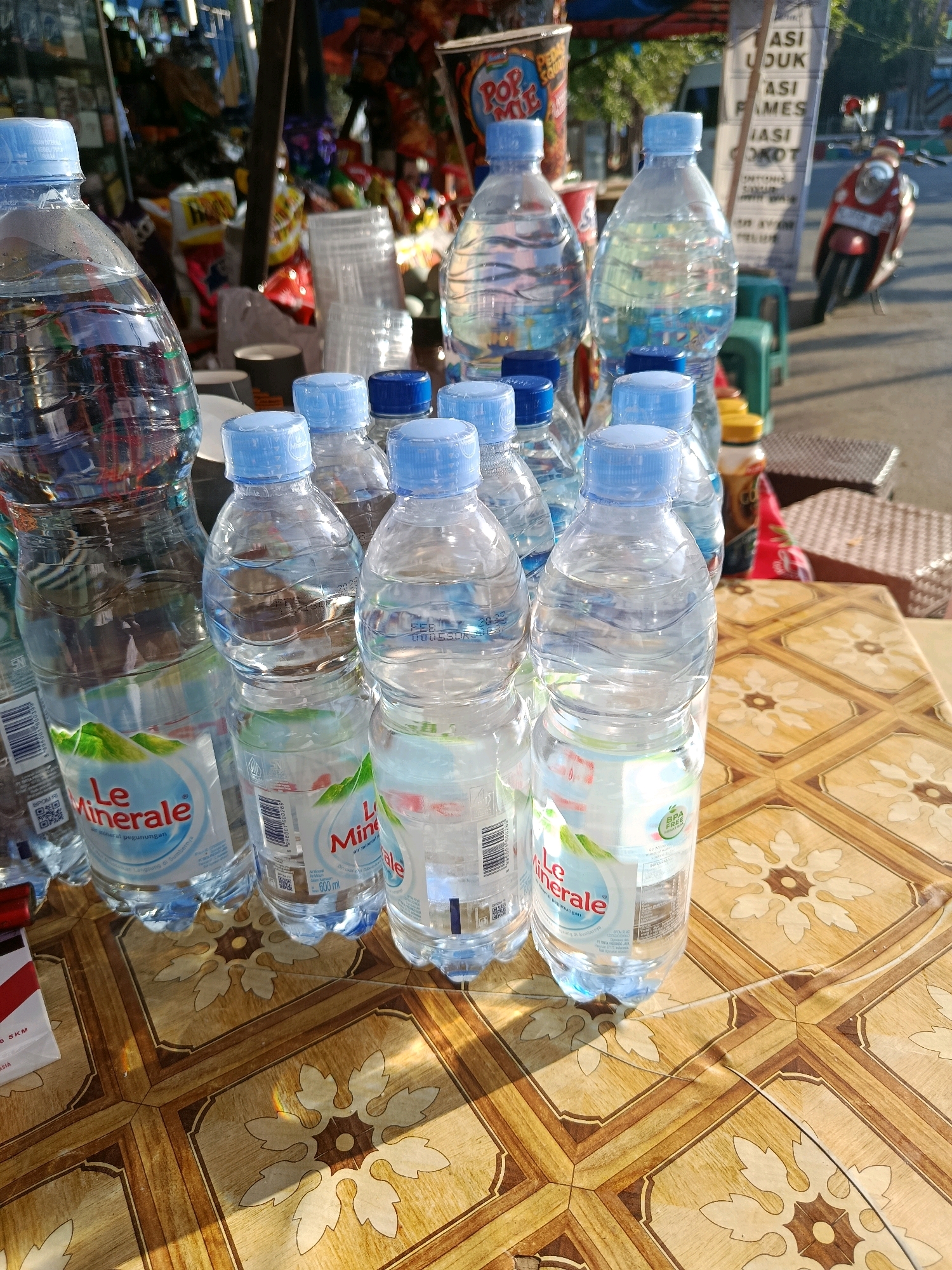 In Jepara, Indonesia, a still life image captures a table laden with bottled water, likely outside a shop. The foreground is dominated by several clear plastic bottles of "Le Minerale" bottled water, each with a light blue cap. The bottles are arranged in a close cluster on a patterned wooden table. Some bottles are partially obscured, and reflections are visible on their surfaces. In the background, shelves are stacked with various goods, including snacks and plastic cups. A scooter, red and white, is parked to the right, and a sign with Indonesian text is visible behind it. The lighting suggests an outdoor setting with sunlight casting shadows.