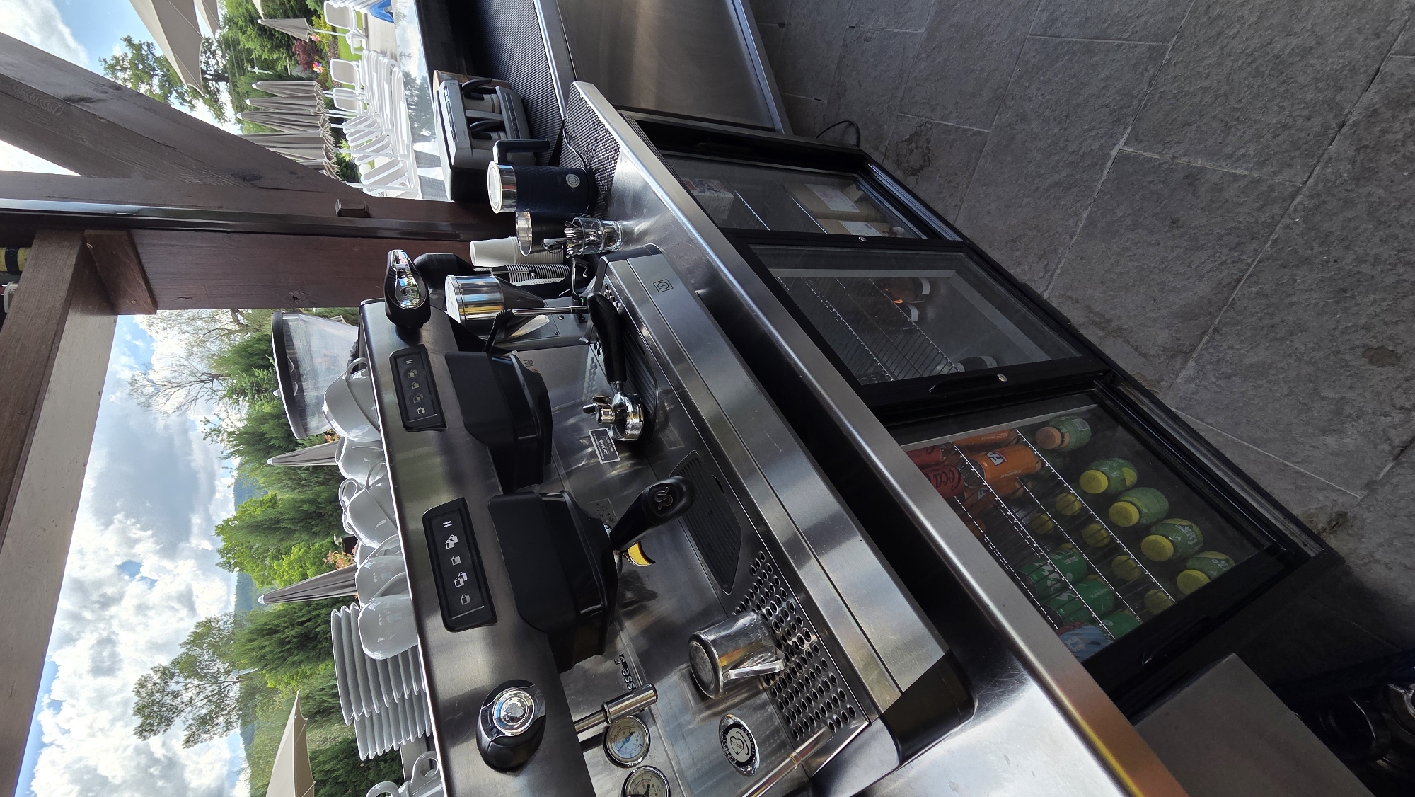 An outdoor coffee bar is seen with a professional espresso machine positioned on a stainless steel counter. Behind the machine, stacked plates and white cups are visible. A small refrigerator with glass doors, filled with bottled beverages, is situated below the counter. In the background, lounge chairs and umbrellas are set up near a body of water. The scene is under a cloudy sky with trees and rolling hills in the distance. The image is taken from a low angle, looking upwards.