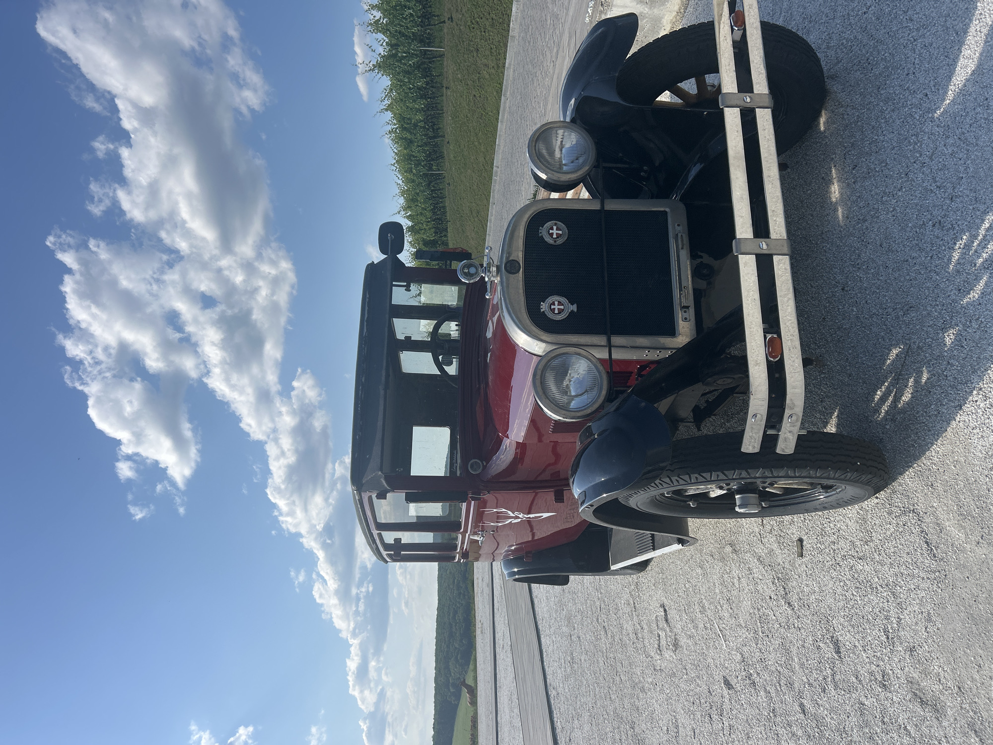 A vintage red automobile with black fenders and roof is parked on a gravel lot. The car has a chrome grille and bumper, two round headlights, and a spare tire on the side. A white deer silhouette emblem is visible on the driver's side door. In the background, a vineyard stretches across a grassy hillside under a bright blue sky with scattered white clouds. To the left of the car, a wooden boardwalk leads towards a distant, tree-lined landscape with a few brown horses grazing. The scene appears to be located in the vicinity of Ulmu, Moldova.