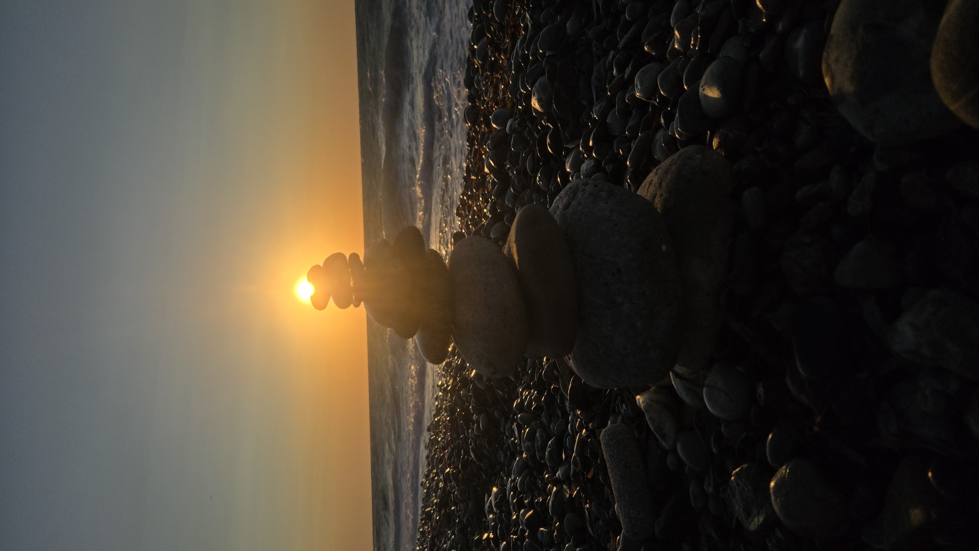 A rock cairn is stacked on a pebble beach with the sun setting over the ocean in the background. The sky is a gradient of orange and blue. The ocean is a deep blue with white waves. The pebbles on the beach are dark and wet. The rock cairn is made of several stacked stones, the largest at the bottom and smaller ones on top. The sun is positioned behind the rock cairn, creating a silhouette effect and lens flare. The scene appears to be peaceful and serene. This photograph was taken at the beach in Soroni, Greece.