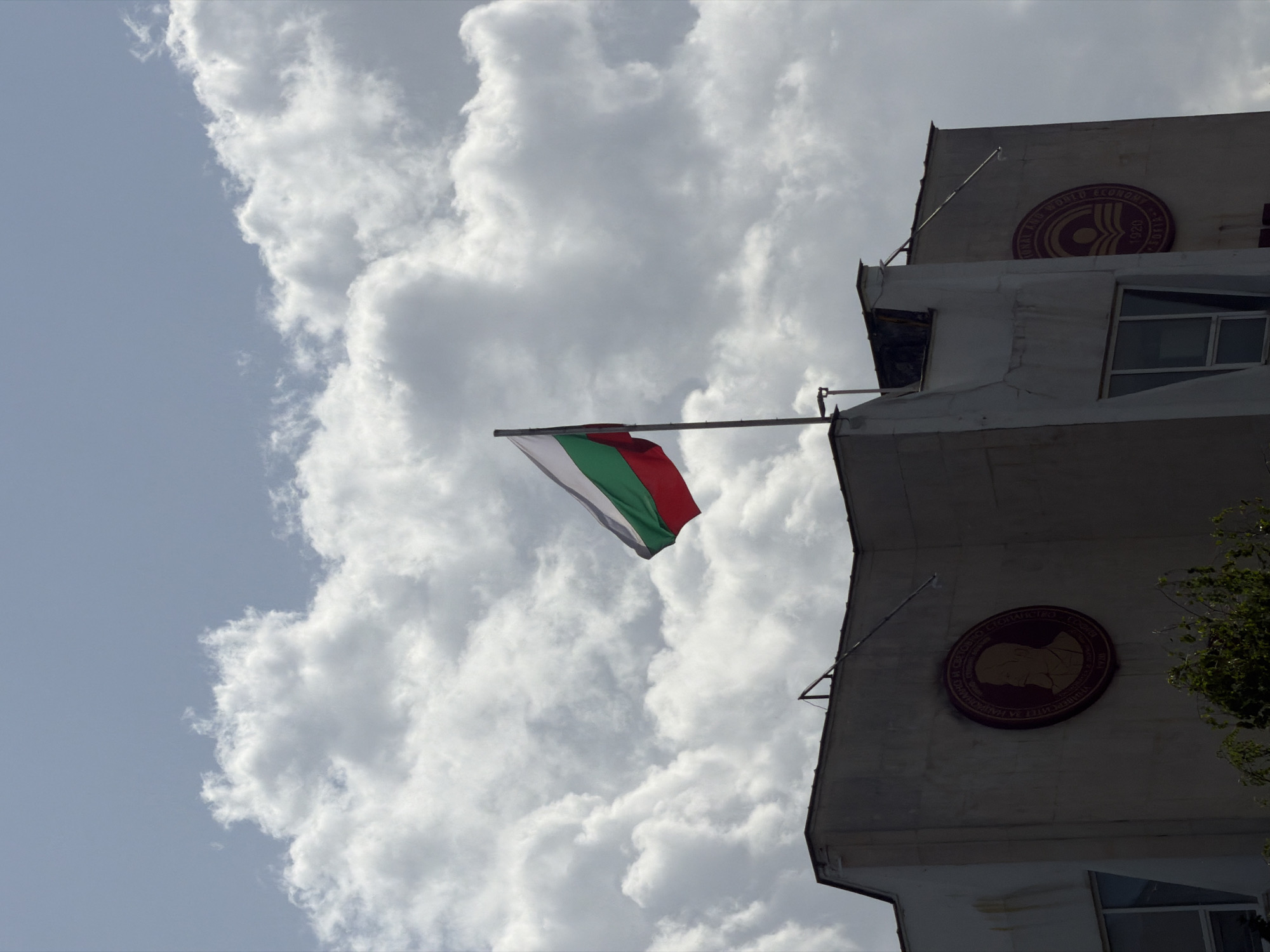 The image shows the Bulgarian flag flying in front of a building in Sofia, Bulgaria. The building appears to be the University of National and World Economy, as indicated by the insignia on the facade. The sky is cloudy.