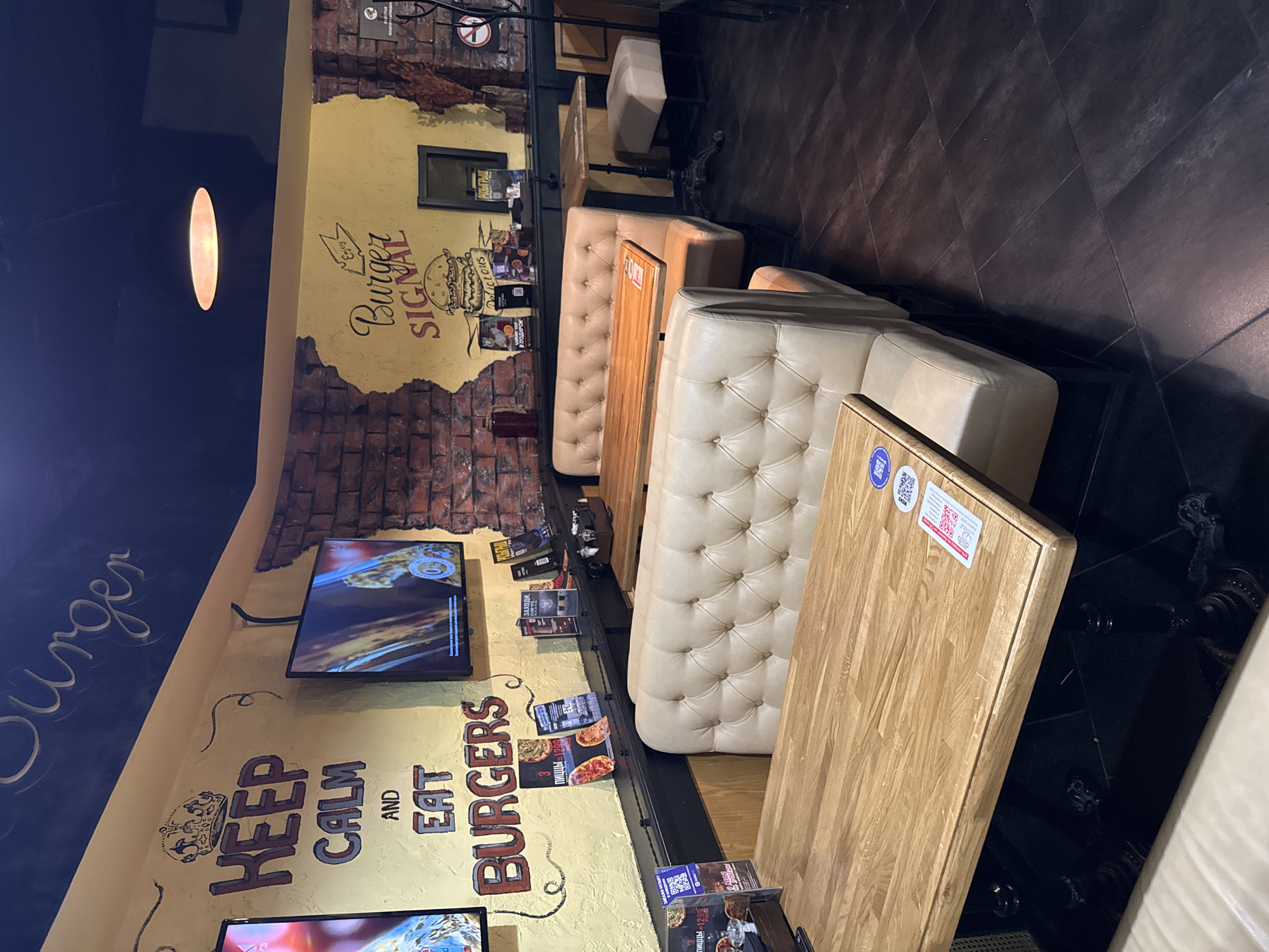 The image captures the interior of a casual burger restaurant, likely located in Russia given the visible Cyrillic text. The scene is devoid of people, offering a static view of the dining area.
The setting features cream-colored, tufted booth seating arranged along one wall with light wooden tables. The walls are predominantly painted yellow, contrasting with sections featuring a faux brick texture. Artificial lighting is provided by a prominent oval-shaped fixture on the dark ceiling. The floor is dark, possibly tiled, and reflective in some areas.
Notable details include extensive wall decorations and signage. A large mural on the wall reads "Burger SIGNAL" alongside a graphic of a burger, with the words "Enjoy" and "Delicious." Further down the wall, another mural features a crown motif and the phrase "Keep CALM AND Eat BURGERS." Multiple flat-screen televisions are mounted on the walls, displaying various content. Menus and advertisements are placed on the tables and along the wall, some featuring QR codes.
Visible text from OCR confirms the establishment's theme and origin: "Burger SIGNAL," "Keep CALM AND Eat BURGERS." Other Russian text includes "3 Пиццы" (3 Pizzas) on a menu, "ЗАХОДИ," (Come in,), "В ПОДАРОК" (As a gift/for free), and "РОЗЫГРЫШ ПИЦЦЫ-1299" (Pizza Raffle-1299), suggesting promotional offers. The overall impression is a modern, themed eatery designed for a casual dining experience.