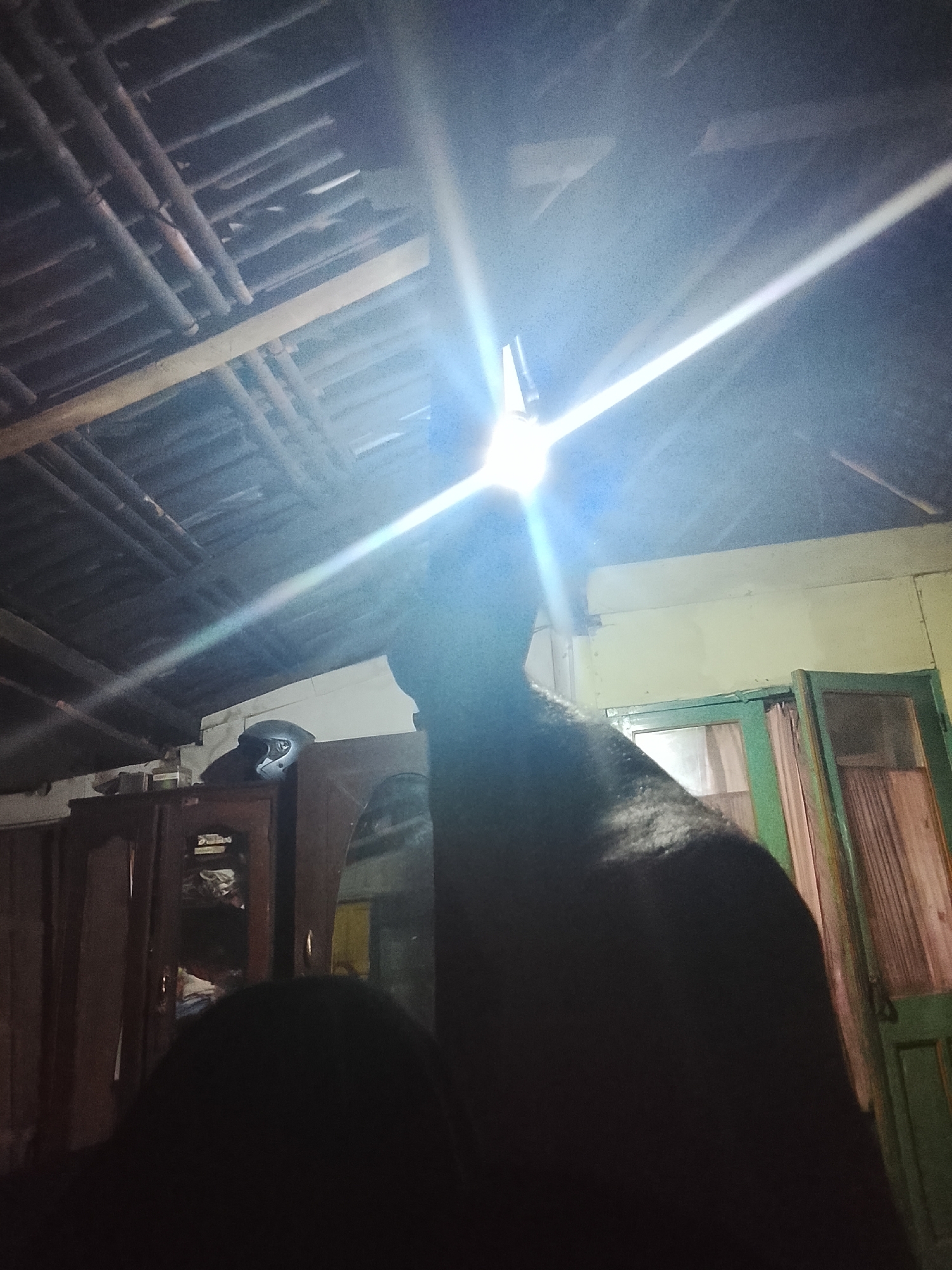 The image depicts an indoor scene, likely a residential room, with a strongly lit ceiling structure composed of wooden beams. A bright, direct light source causes significant lens flare and glare, obscuring details in the upper center of the frame. In the foreground and mid-ground, a dark, silhouetted figure occupies a significant portion of the image. To the left, a dark wooden cabinet with shelves is partially visible, and on top of it sits a white motorcycle helmet. To the right, a green-framed door with wooden panels and a curtain is present. The overall environment suggests a simple, possibly rustic interior. The image is described as being taken in Mernek, Indonesia, though this specific location is not visually identifiable from the image content itself.