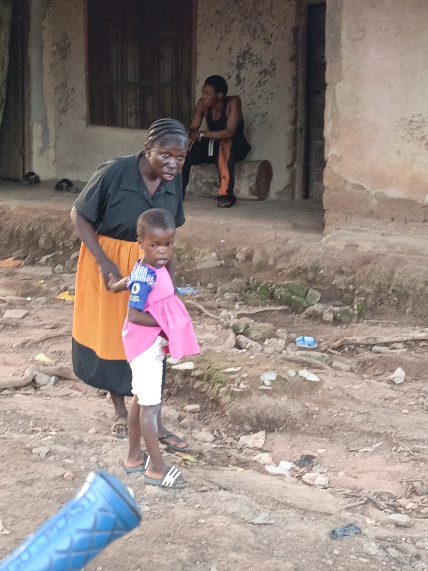 In Akwanga, Nigeria, a woman wearing a black shirt and an orange skirt stands on a dirt path, holding the hand of a young child. The child is dressed in a pink dress with blue accents and white shorts. In the background, another woman is seated on a log, wearing a black tank top and orange-striped pants. The environment appears to be an outdoor area with a rustic building and debris scattered on the ground.