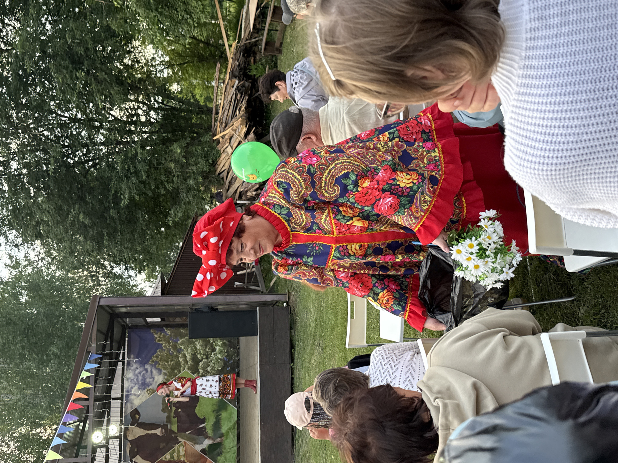 An outdoor event is underway in the village of Kukui, Russia, where a woman in a colorful traditional Russian costume, adorned with red and white polka dots on her headscarf, is presenting a bouquet of daisies to an unseen recipient. She is holding the flowers and a black garbage bag. The woman is standing in front of an audience seated in white chairs on a grassy area. Behind the audience, a stage is set up with a backdrop featuring an image of cows and trees, where another woman, dressed in a similar style, is singing into a microphone. The background of the scene includes trees, a pile of wood, and a green balloon floating in the air. The overall atmosphere appears to be a festive community gathering or performance.