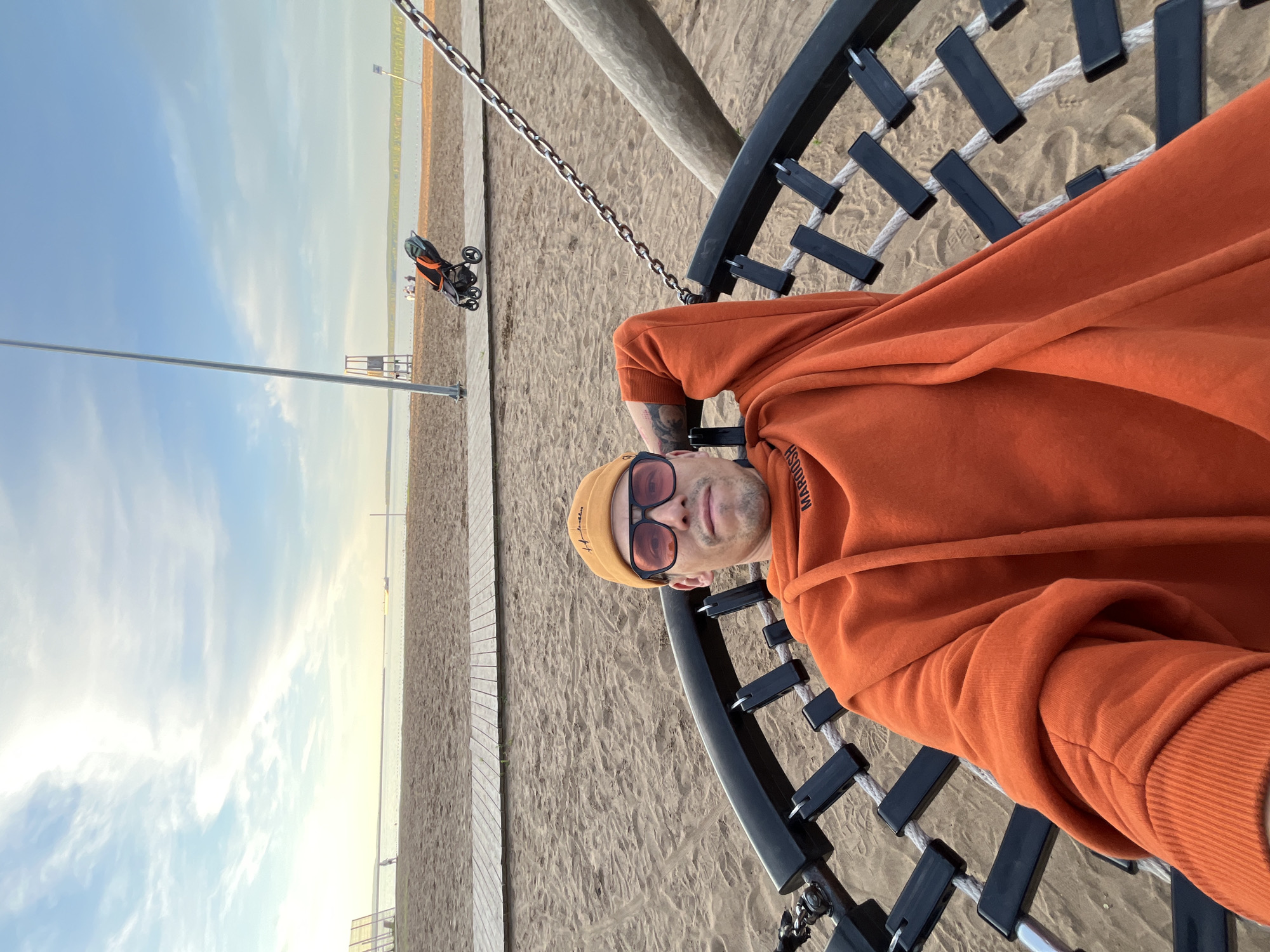 The image shows a man taking a selfie at a sandy beach or lakeside recreational area, likely in Chaikovsky, Russia. The man is positioned in the foreground, looking directly at the camera. He is wearing a bright orange hoodie, a yellow beanie, and sunglasses with distinct orange-tinted lenses. The word "MAROOSH" is partially visible on his hoodie, and "Helsinki" or "Helsinky" can be seen on his beanie. He is sitting on a large, circular swing made of black metal and ropes, which partially frames him.
The background features a vast sandy area, suggesting a beach, with a wooden boardwalk or path running alongside it on the left. Beyond the sand, a calm body of water stretches towards the horizon, meeting a clear sky with scattered, wispy clouds. The lighting indicates late afternoon or early evening, with a gentle, warm glow illuminating the scene, particularly over the water. In the mid-ground, a black and orange baby stroller is visible on the sand. A tall, slender metal pole stands vertically on the left side of the frame, near the boardwalk. The scene suggests a tranquil, leisure setting by the water.