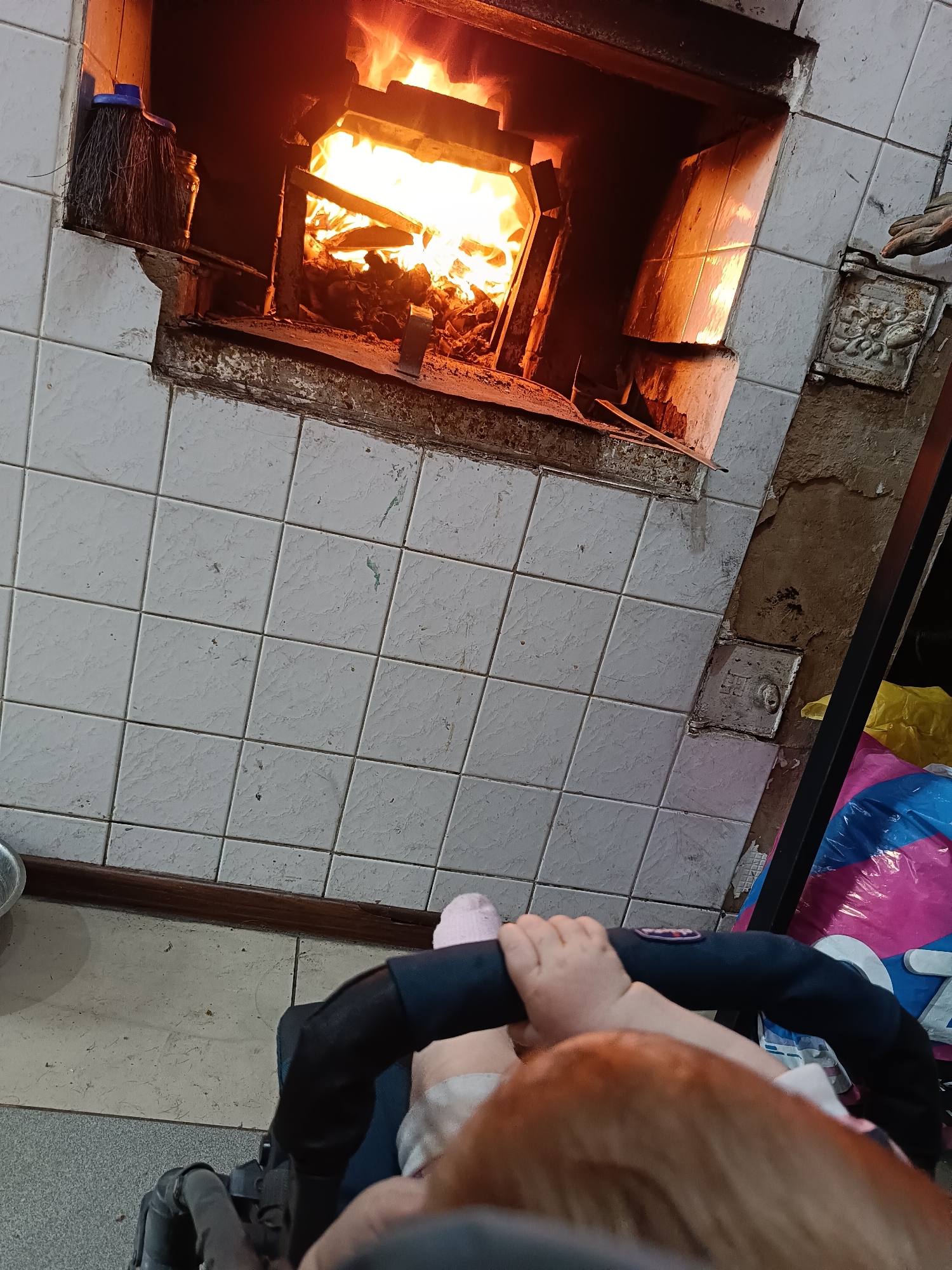 A low-angle shot depicts a fire burning in a tiled oven, with a child visible in the foreground. The oven is surrounded by white tiles with a brown and damaged border. A blue broom leans against the left side of the oven. A metal plate with a design is to the right of the oven. In the lower part of the image, a child in a stroller is visible, with their hands gripping the black stroller bar and their pink socked foot visible. The baby's red hair can be seen. Beside the stroller, there is a colorful bag.