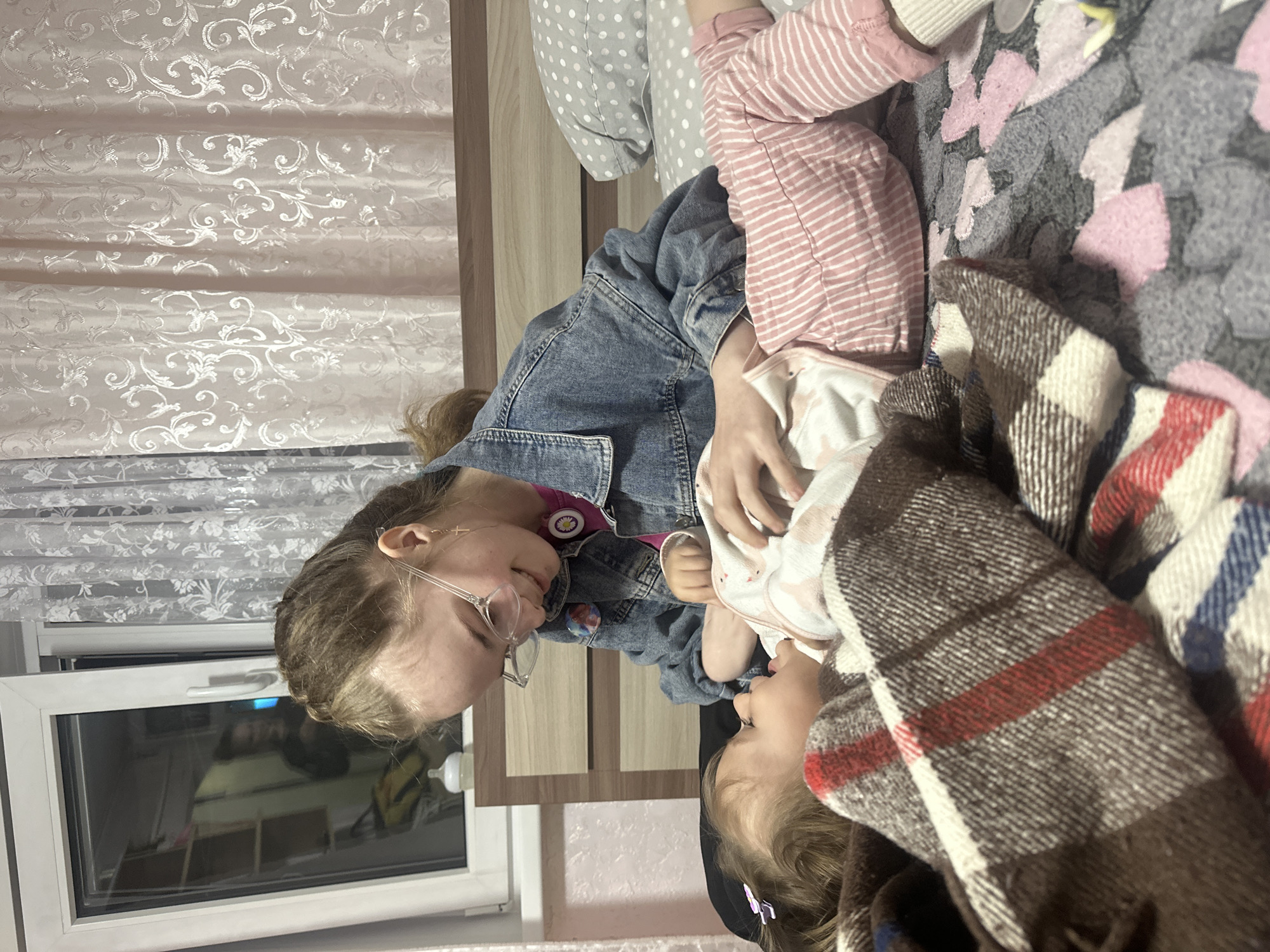 Two Caucasian females are interacting on a bed in an indoor setting. The older female, wearing glasses and a denim jacket, is facing and gazing at a younger female who is partially covered by a brown, white, and red plaid blanket. The younger female is lying down. The bed has a patterned grey and pink blanket, and the person above the older female is wearing pink and white striped pants and is lying on a grey pillow with white dots. A window with white blinds is visible. The location is Uva, Russia.