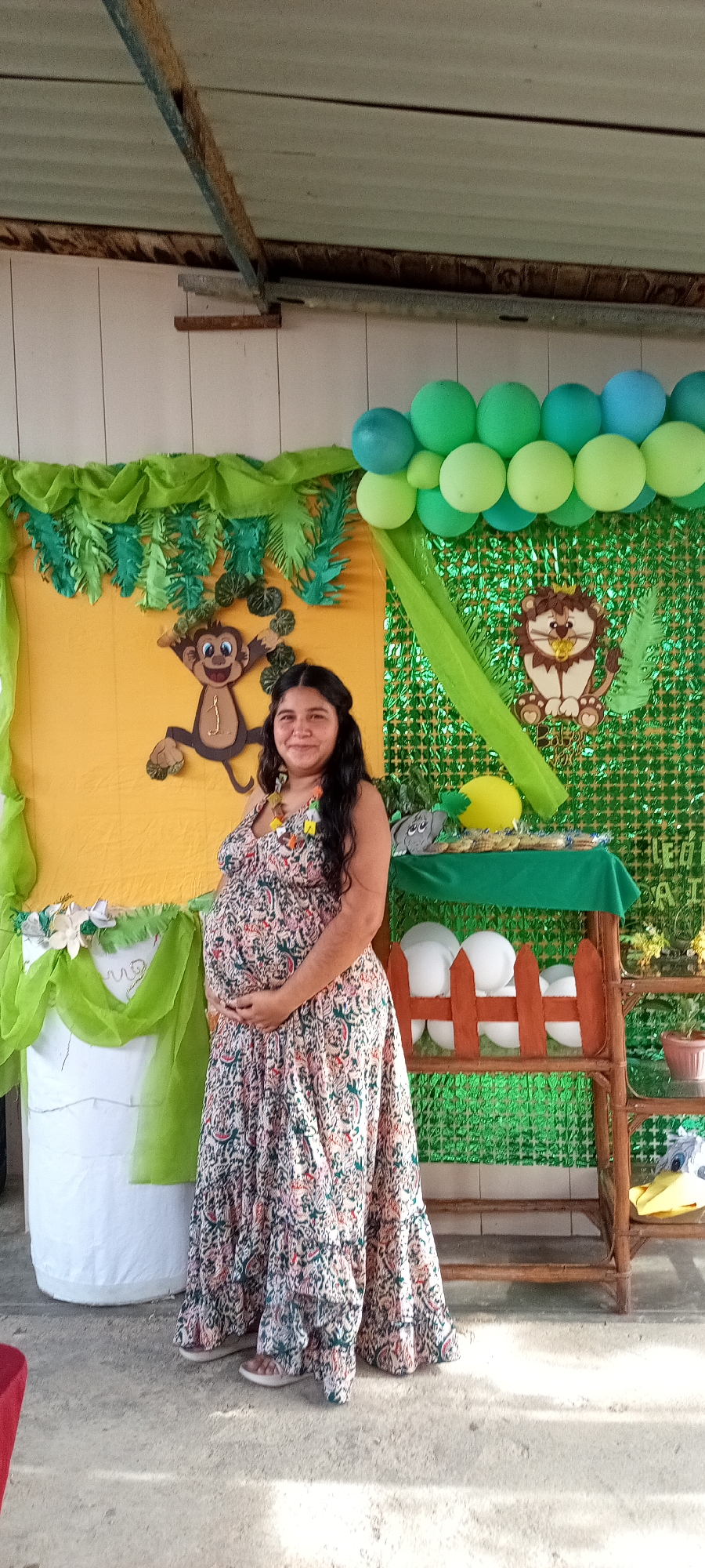 Here is a description of the image:
A pregnant woman stands smiling in front of decorations, likely at a baby shower or similar event. The background is decorated with yellow and green colors, featuring cartoon images of a monkey and a lion. There are green and blue balloons arranged above the decorations. A small wooden shelf sits to the right of the woman, with various items displayed on it. The woman wears a patterned maxi dress and has her hands placed on her baby bump. The location appears to be outdoors or in a semi-outdoor space.