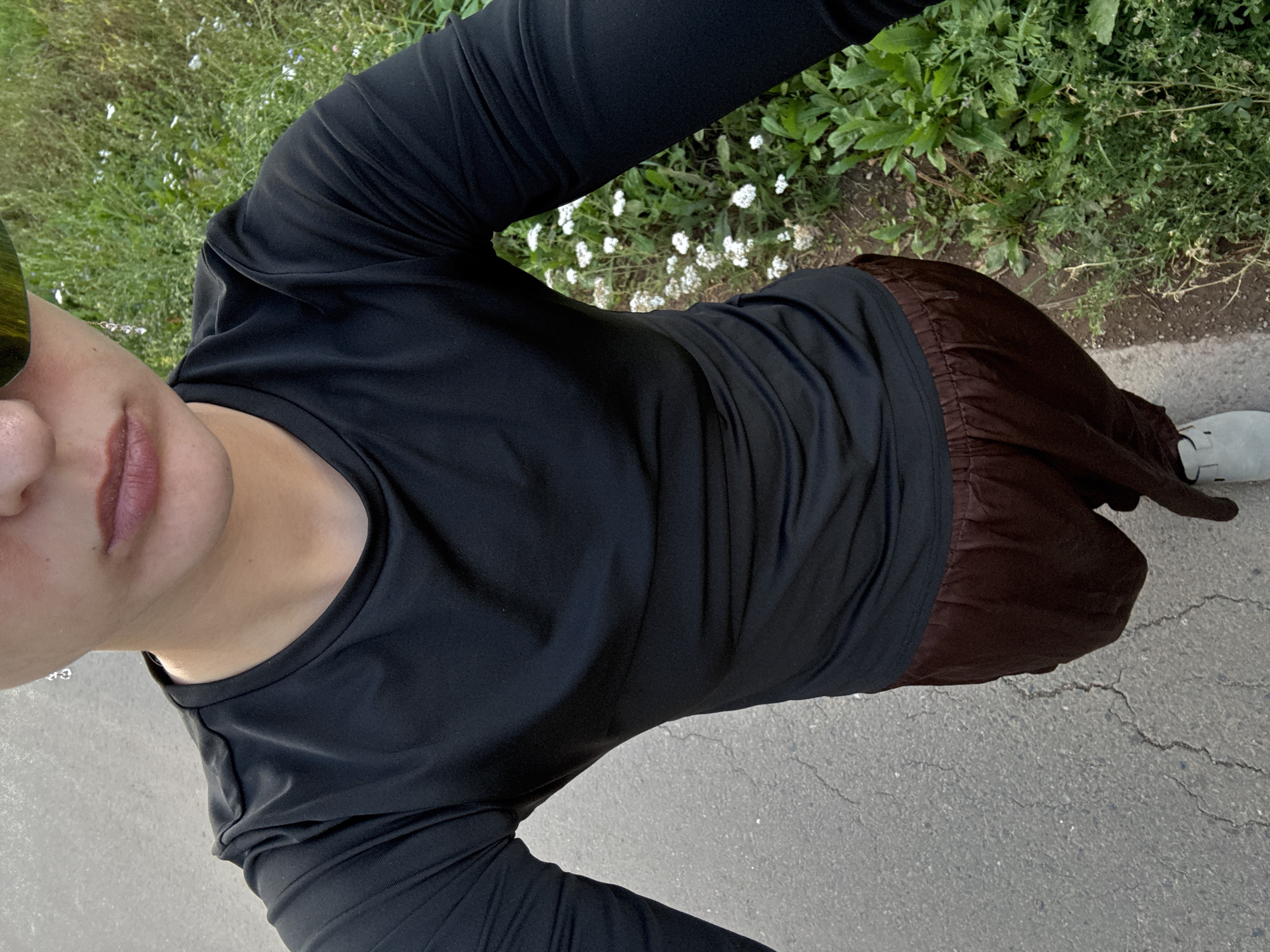 A person is captured from a high angle, looking down at their own torso and lower body. They are wearing a black long-sleeved shirt and brown pants. The person is standing on a paved surface, possibly a path or sidewalk, with lush green foliage and small white flowers to their right. The lighting suggests it is daytime. The background appears to be an outdoor natural setting with vegetation. The image does not contain any elements that indicate a specific location within the city of Подшивалово, Russia.