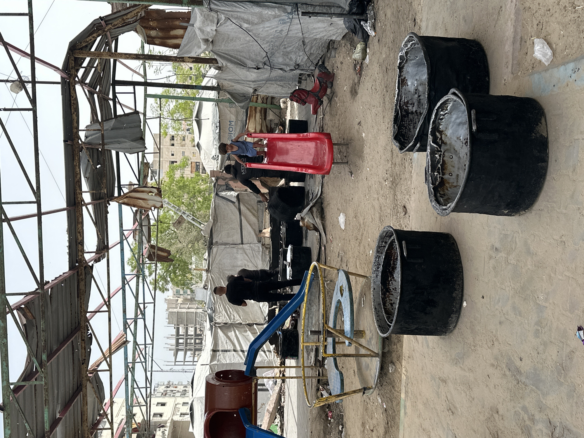 The image shows a playground in Gaza City, Palestinian Territories. The playground is in disrepair, with damaged metal structures and tents erected nearby. Children are playing on a red slide. Adults are present. There are several black, cylindrical containers. Buildings are visible in the background. The environment suggests poverty and displacement.