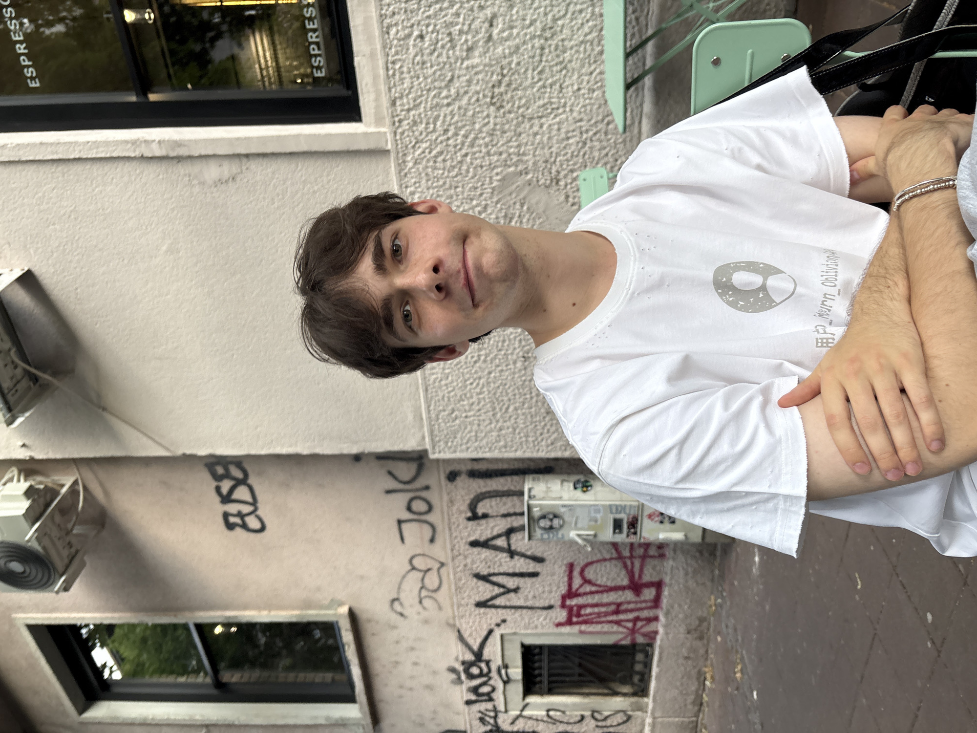 A young man with brown hair and brown eyes is seated outdoors, facing the camera. He is wearing a white t-shirt with a circular grey graphic and some text. His arms are crossed. Behind him, graffiti covers the wall of a building. The graffiti includes the words "MADI" and "Joki". There is also a window with "ESPRESSO" written on it. A green metal chair and table are visible to the right of the man. The ground is paved with reddish-brown bricks. The overall setting appears to be a street in Belgrade, Serbia, likely near a cafe.