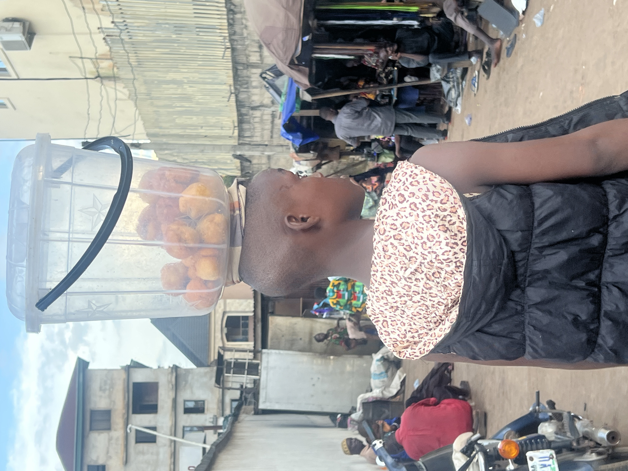 A young person, viewed from behind and in profile, walks forward with a clear plastic bucket full of fried dough balls balanced on their head. The individual wears a black puffer vest over a patterned top. The background depicts a bustling marketplace in Nkpor, Nigeria, with various stalls, people, and buildings. A motorcycle is visible to the left, and a weathered fence lines the right side of the frame. The sky above is overcast.