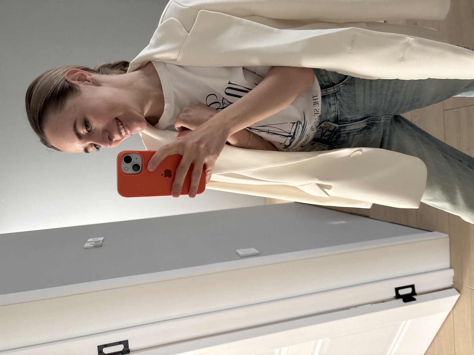 A young woman smiles at an orange iPhone as she takes a selfie indoors. She wears blue jeans, a white t-shirt with a boat graphic, and a white blazer. The background shows a white door with a black handle and light gray walls. The location is not mentioned in the image.