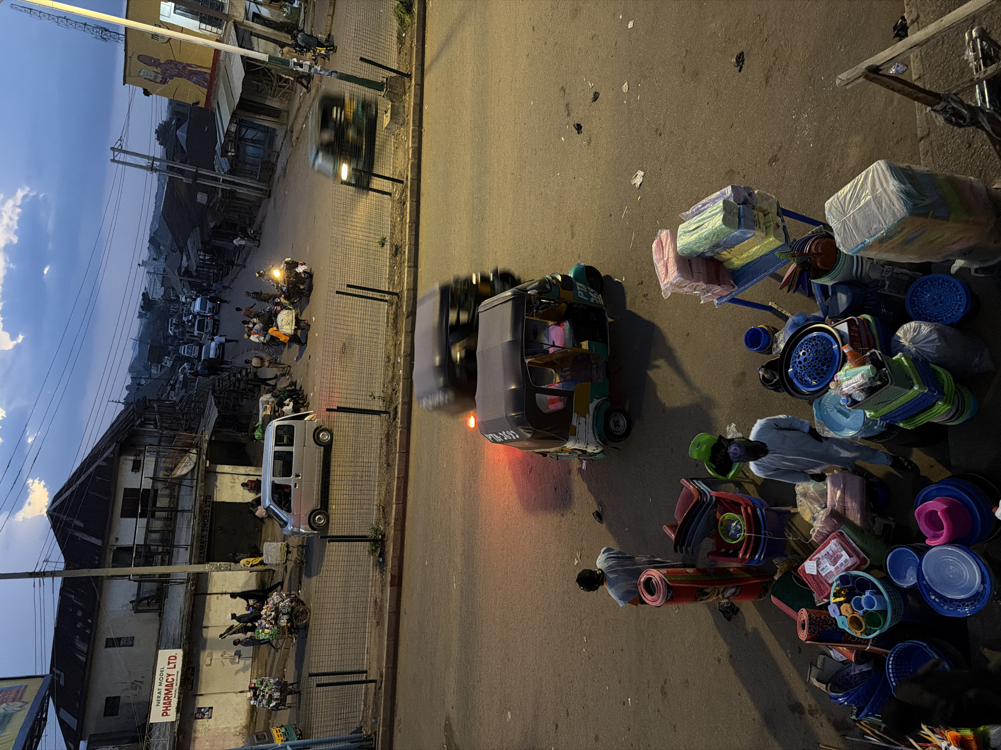 The image depicts a street scene in Bukuru, Nigeria, at dusk. A street vendor's stall is in the foreground, displaying various household items such as plastic basins, buckets, chairs, and rolled-up mats. Two individuals, dressed in traditional Nigerian attire, are visible near the stall. One is standing by the items for sale, and the other is walking away. A three-wheeled auto-rickshaw (Keke Napep) is parked on the right side of the street, its brake lights illuminated. Further down the street, other vehicles, including cars and more auto-rickshaws, are present, some in motion. Buildings line the street, some appearing to be shops with signage, one of which reads "NERAT MODEL PHARMACY LTD." The sky is overcast with hints of a twilight glow. The overall atmosphere suggests a busy commercial area during a transitionary period from day to night.