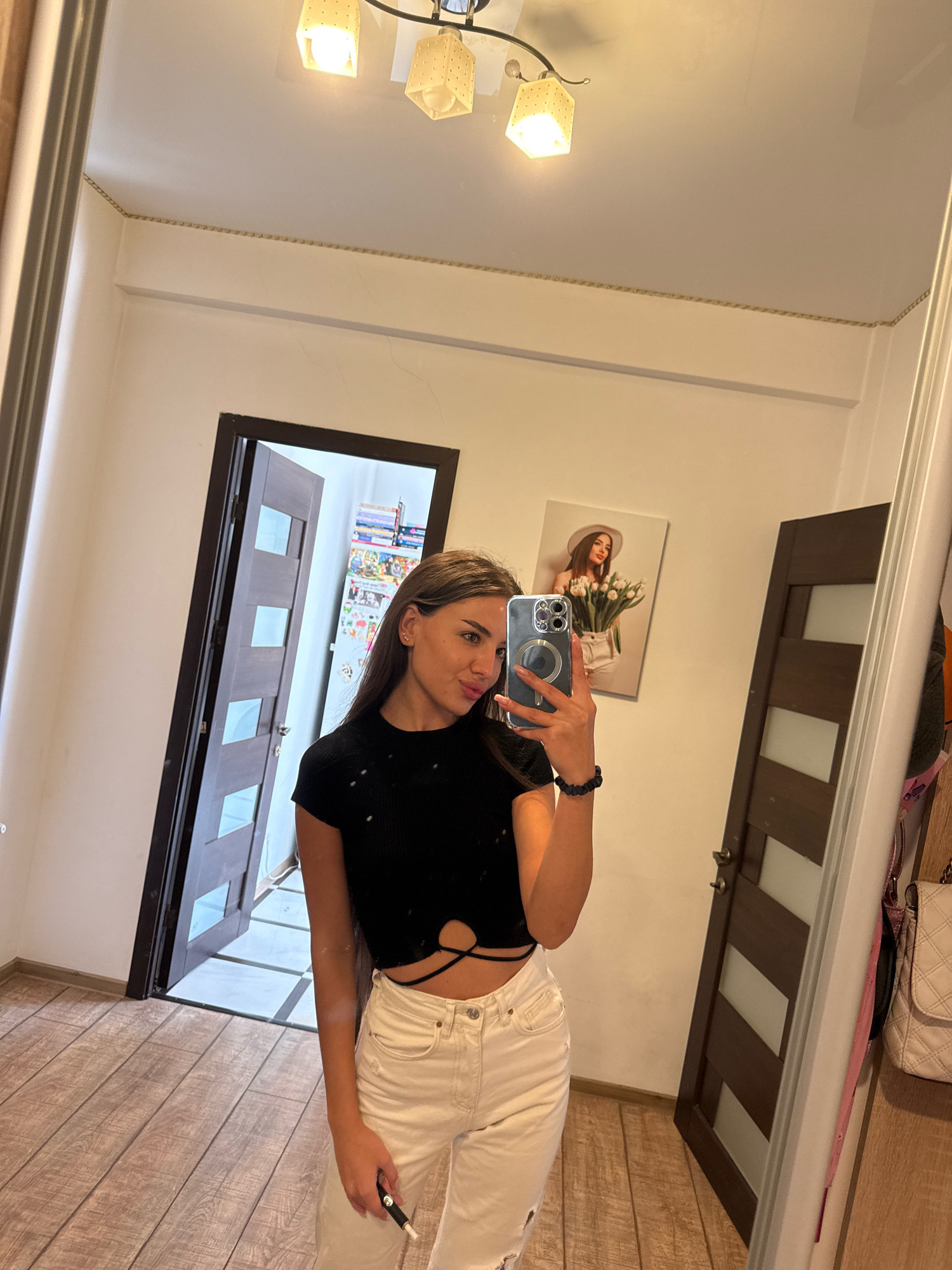 The image shows a young woman taking a selfie in a room, possibly her home, in Chișinău, Moldova. She is wearing a black crop top with cutouts and white ripped jeans. She is holding a smartphone to take the picture. In the background, there is a large mirror reflecting the room, which appears to be a hallway or entryway. There are two doors visible, one slightly ajar, revealing shelves filled with books and other items. A framed picture of a woman holding a bouquet of flowers hangs on the wall. The flooring is made of light-colored wooden planks. The overall lighting suggests it is daytime.