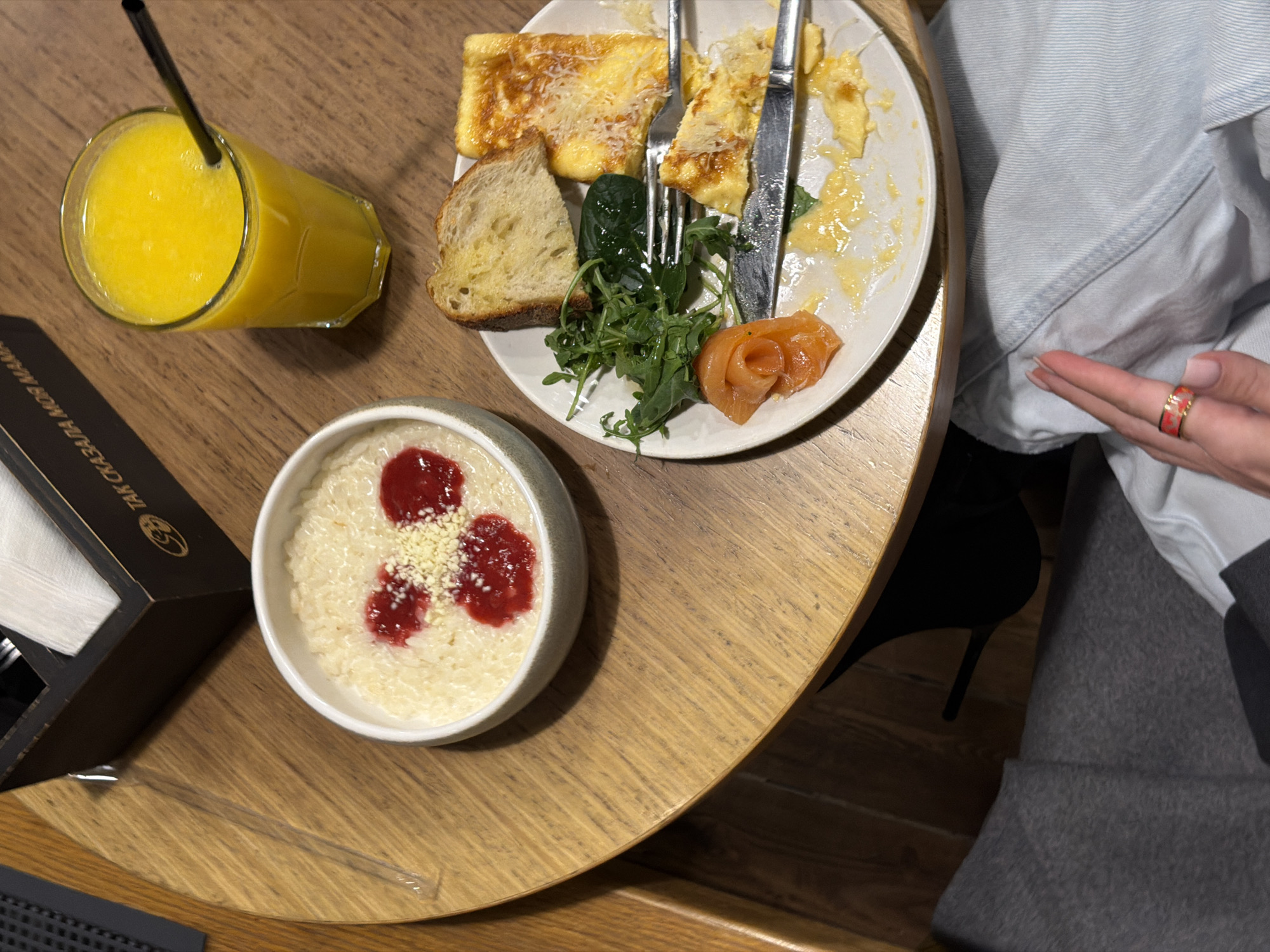 The image depicts a meal served on a light wooden table in a "Tak Skazala Moya Mama" restaurant in Moscow, Russia. In the upper left, a dark wooden napkin dispenser with white napkins is partially visible, bearing the restaurant's name in Russian script. To its right, a clear glass holds a vibrant yellow beverage with a black straw. A white bowl in the center contains a creamy white dish, likely porridge, garnished with three dollops of red jam and a sprinkle of light-colored crumbs. A white plate, positioned on the right, shows a partially consumed meal consisting of a piece of toasted bread, a portion of cooked egg topped with grated cheese, a small cluster of green leaves (arugula and spinach), and a rolled slice of salmon. A fork and knife rest on the plate. The lower part of the frame reveals the lower legs of a person, with one hand featuring manicured nails and a red-and-gold ring, resting on light blue denim fabric.