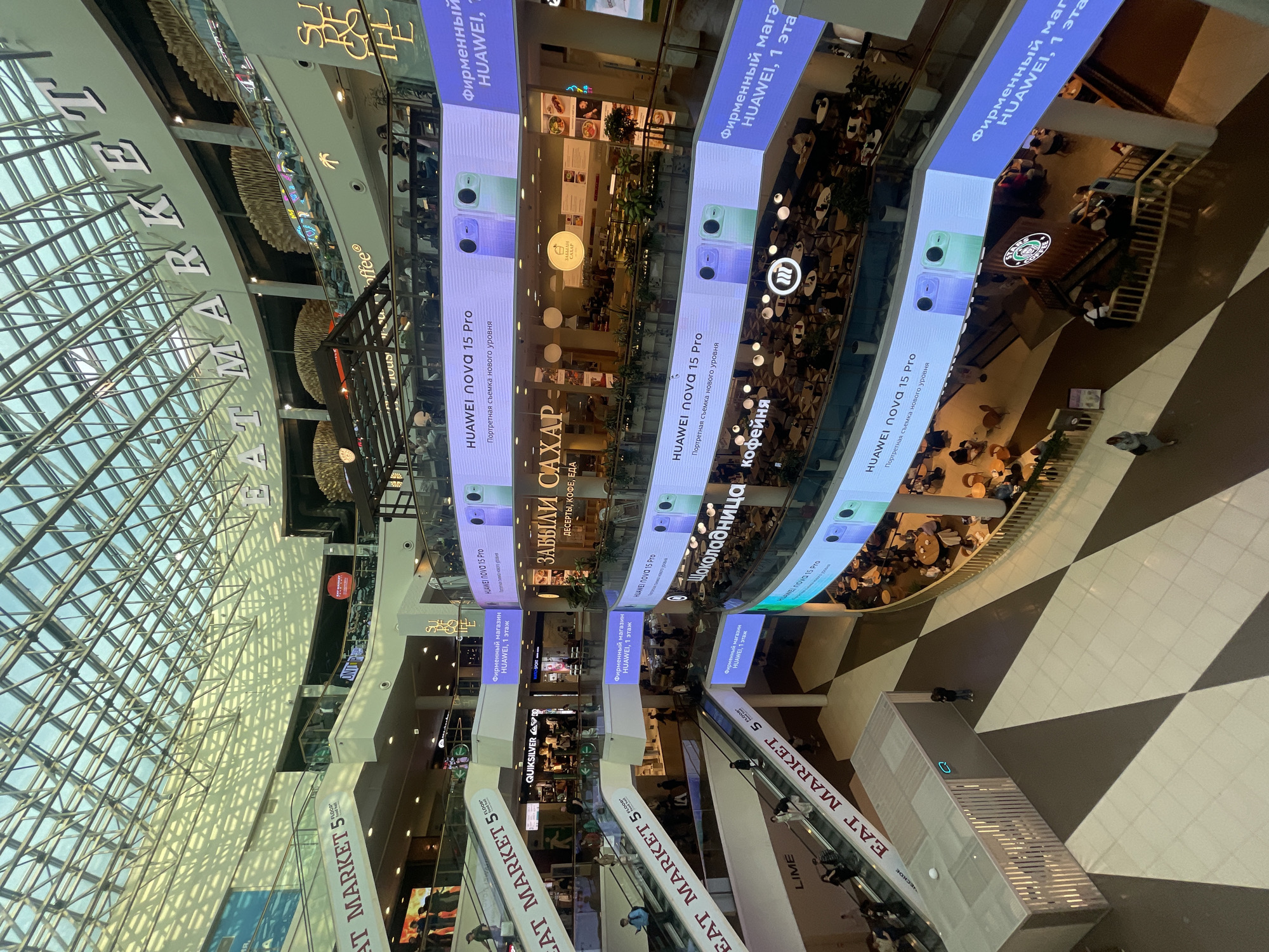 The image captures an interior view of a large shopping mall, likely in Saint Petersburg, Russia, as suggested by the Cyrillic text on the signage. The scene is taken from an elevated perspective looking down into the mall's atrium.
The mall features multiple levels visible due to the open design. Banners advertising "HUAWEI nova 15 Pro" are suspended from the ceiling. There are advertisements for various restaurants and stores including "ЗАБЫЛИ САХАР" (Zabyli Sahar), "Шоколадница кофейня" (Shokoladnitsa Coffee), and "EAT MARKET 5".
Escalators are visible, labeled "EAT MARKET 5 FLOOR". People are observed walking on various levels of the mall and riding the escalators. The mall has a large, glass-covered roof providing natural light. The overall architecture suggests a modern design with a mix of retail and dining establishments.