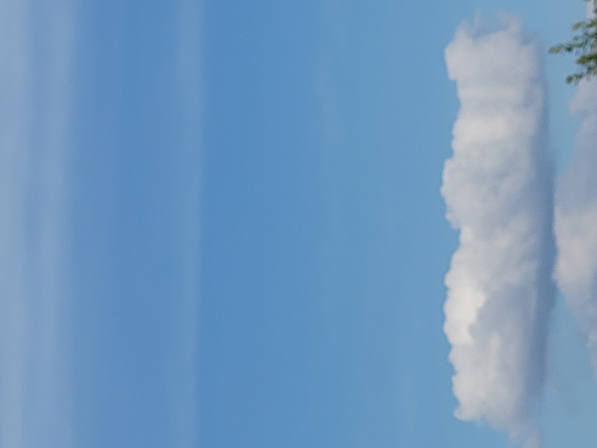 The image depicts a clear blue sky with a few fluffy white clouds concentrated in the lower portion of the frame. In the bottom right corner, a small section of green foliage from a tree is visible, suggesting an outdoor scene. The overall impression is that of a bright, daytime sky.