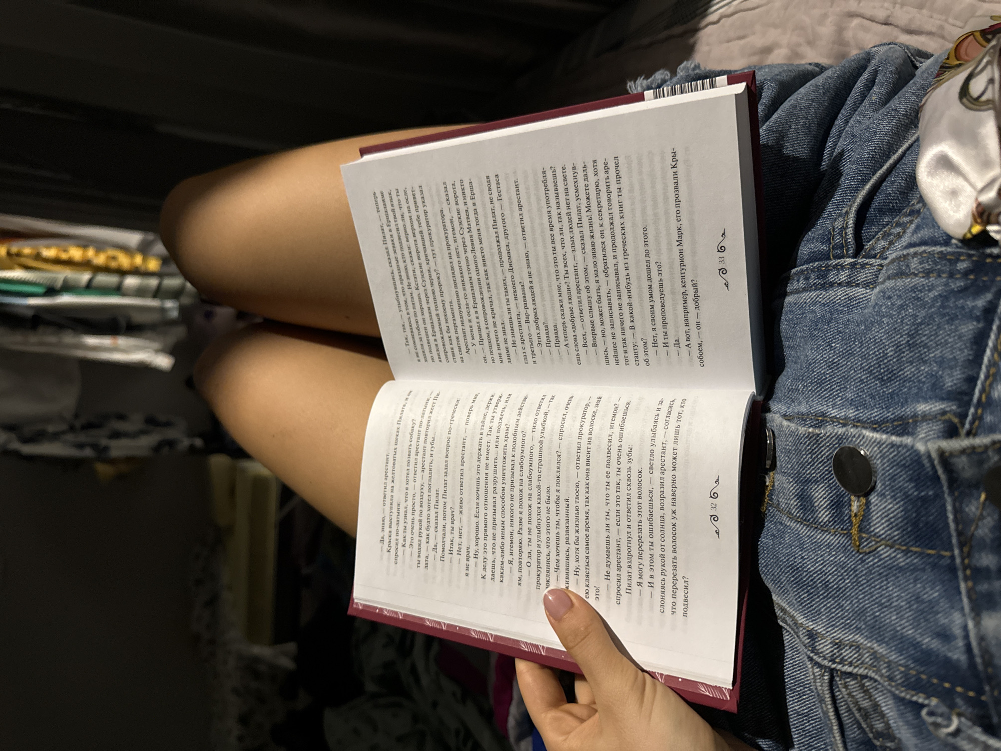 A person is sitting with a book open in their lap. The book has a red cover and the pages are filled with text in a Cyrillic script. The person's hand is visible, holding the book open. They are wearing denim shorts. The setting appears to be indoors, possibly in a bedroom or living area, as suggested by the presence of bedding or cushions.