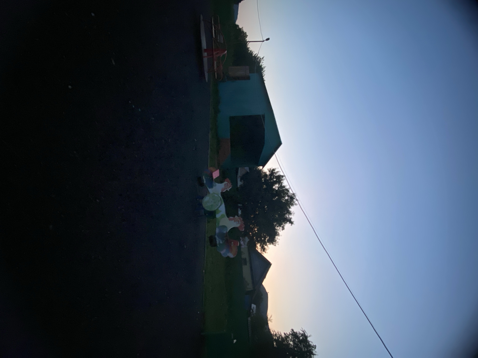 The image captures an outdoor scene at dusk in Maloarslangulovo, Russia, featuring a playground and surrounding structures. The sky is a gradient of clear blue at the top, transitioning to lighter hues near the horizon where a subtle sunset is visible. A power line stretches diagonally across the upper portion of the frame. In the mid-ground, a teal-colored, barn-like structure with a large, dark opening dominates the left side. To its left, a red merry-go-round stands on a dark, asphalt-like surface. A multi-colored rocking toy, resembling two roosters, is positioned to the right of the barn. Behind these playground elements, several houses with peaked roofs and trees are visible against the fading light. The foreground consists of the same dark ground surface. No people are present in the scene.