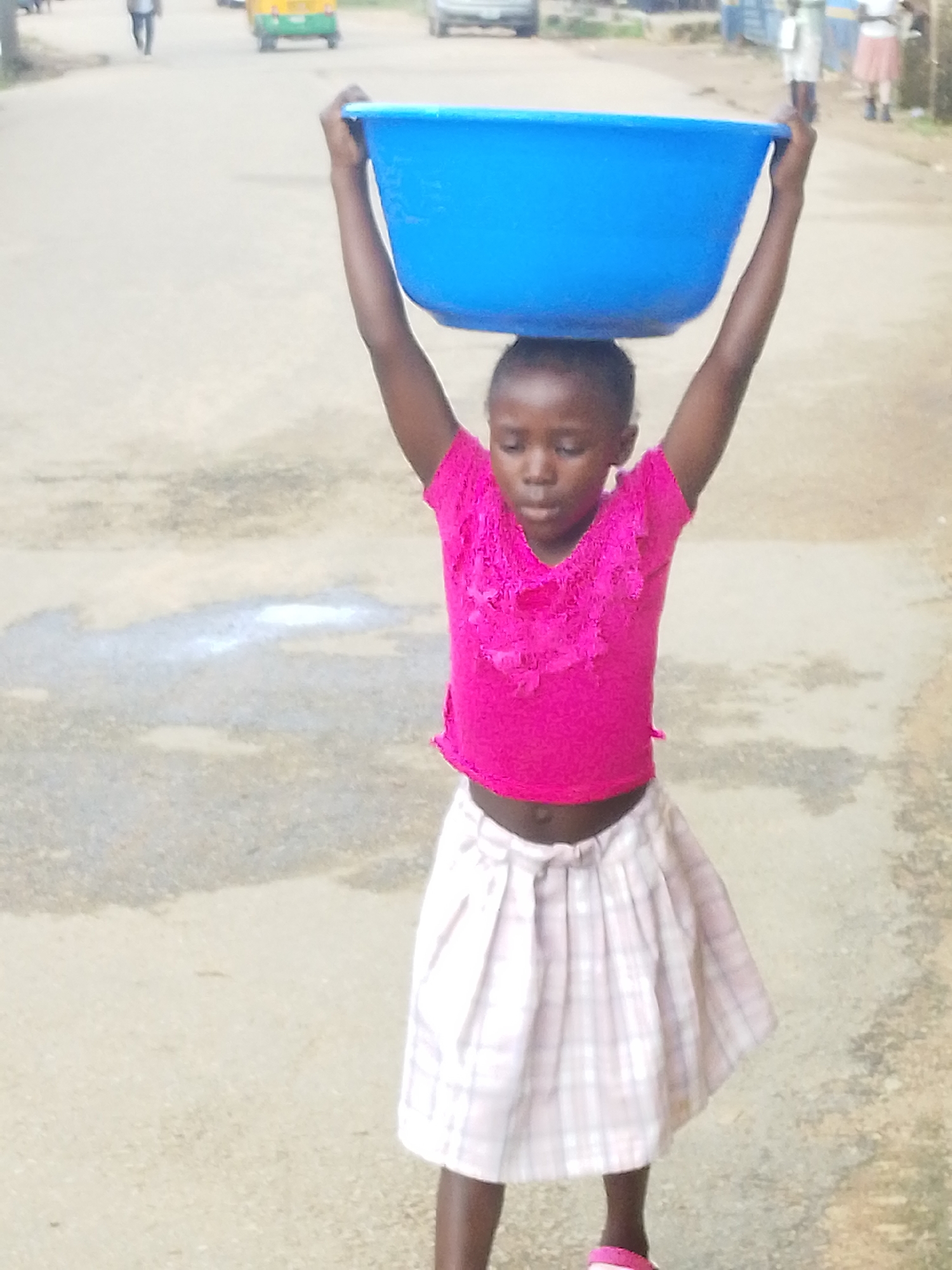 In Jos, Nigeria, a young girl carries a large blue basin on her head while walking on a paved road. She is wearing a bright pink ruffled top and a white and pink plaid skirt. Her arms are raised to balance the basin. In the background, other people and vehicles are visible, including a yellow and green auto-rickshaw, suggesting a busy urban environment. The road surface shows signs of wear and some scattered water or debris.