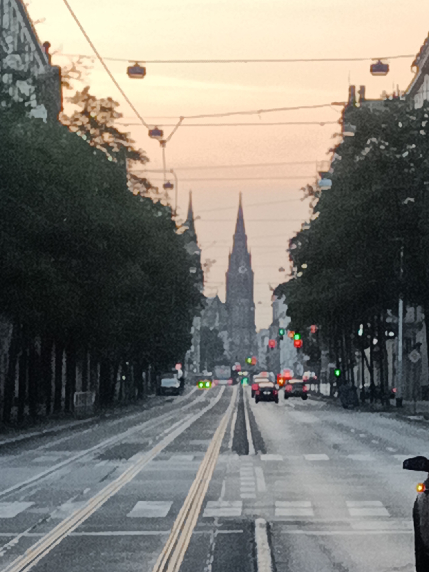 An eye-level medium shot captures a street scene in Prague, Czechia, during twilight or early morning. A streetcar track runs through the center of the frame, with yellow markings indicating its path. On either side of the street, tall, dark trees line the sidewalks, obscuring most of the buildings behind them. In the distance, the iconic spires of St. Vitus Cathedral rise above the cityscape, silhouetted against a pale orange and pink sky. Several cars are visible on the road, their headlights and taillights illuminated. Traffic lights, some green and some red, are present at intersections. Overhead, a series of cable car pods are suspended from wires, adding another layer to the urban infrastructure. The overall mood is serene and evocative, with the fading light casting a soft glow over the city.