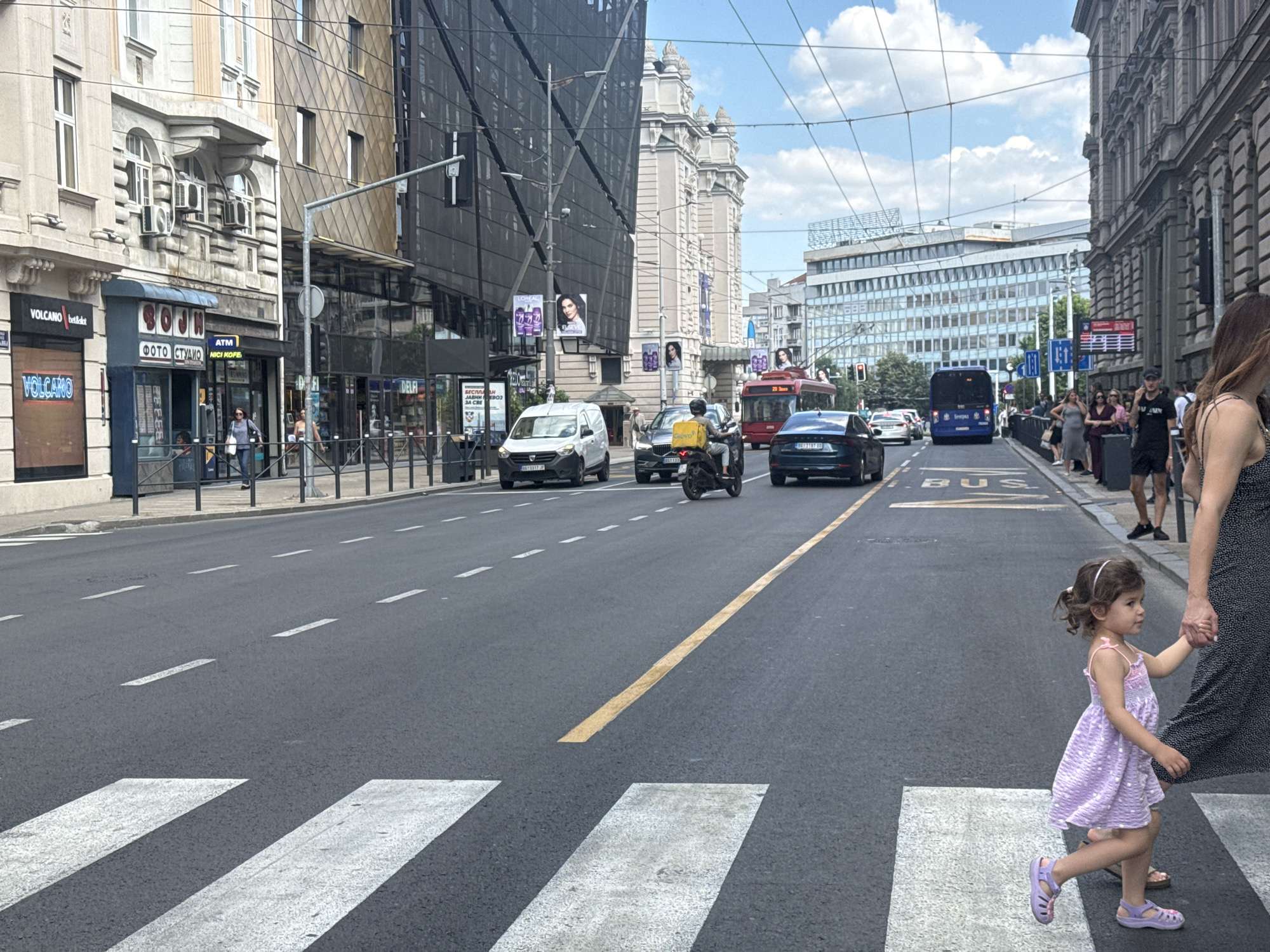 A daytime street scene in Belgrade, Serbia, shows a wide asphalt road with painted white lines for a pedestrian crossing in the foreground. A young girl in a purple polka-dot dress and sandals, holding an adult's hand, is walking across the crosswalk. To the left of the street, a row of buildings lines the sidewalk. One building features storefronts with signage including "VOLCANO," "bet&set," "SOJN," "ФОТО СТУДИО," "ATM," and "МІСІ КОFE." Another building has a dark, modern facade covered in a black mesh material, with posters of a woman's face and "LOREAL" branding. Further down the street, a white van, a motorcycle with a delivery rider, and cars are visible. A red bus is also on the road, and blue signage with "BUS" is painted on the asphalt. The sky above is partly cloudy, suggesting fair weather. In the background, more buildings of varying architectural styles can be seen, including a large, light-colored classical building and a modern glass-fronted building. The overall impression is a busy urban environment on a bright day.