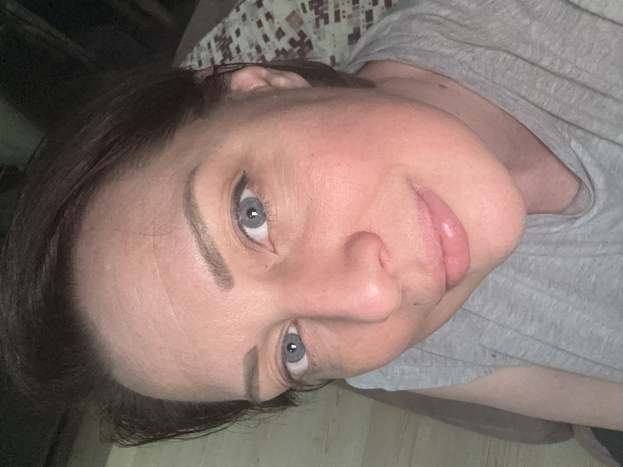 The image shows a close-up selfie of a woman's face, taken from a slightly high angle. She has short, dark brown hair, fair skin with some redness on her cheeks, and light blue eyes. Her eyebrows are well-defined and dark. She is wearing a grey t-shirt. Her lips are slightly parted and appear to have a subtle gloss. The background is blurred, but suggests an indoor setting with patterned fabric behind her. There is no specific indication of the city of Gölbaşı, Turkey, within the image itself.
