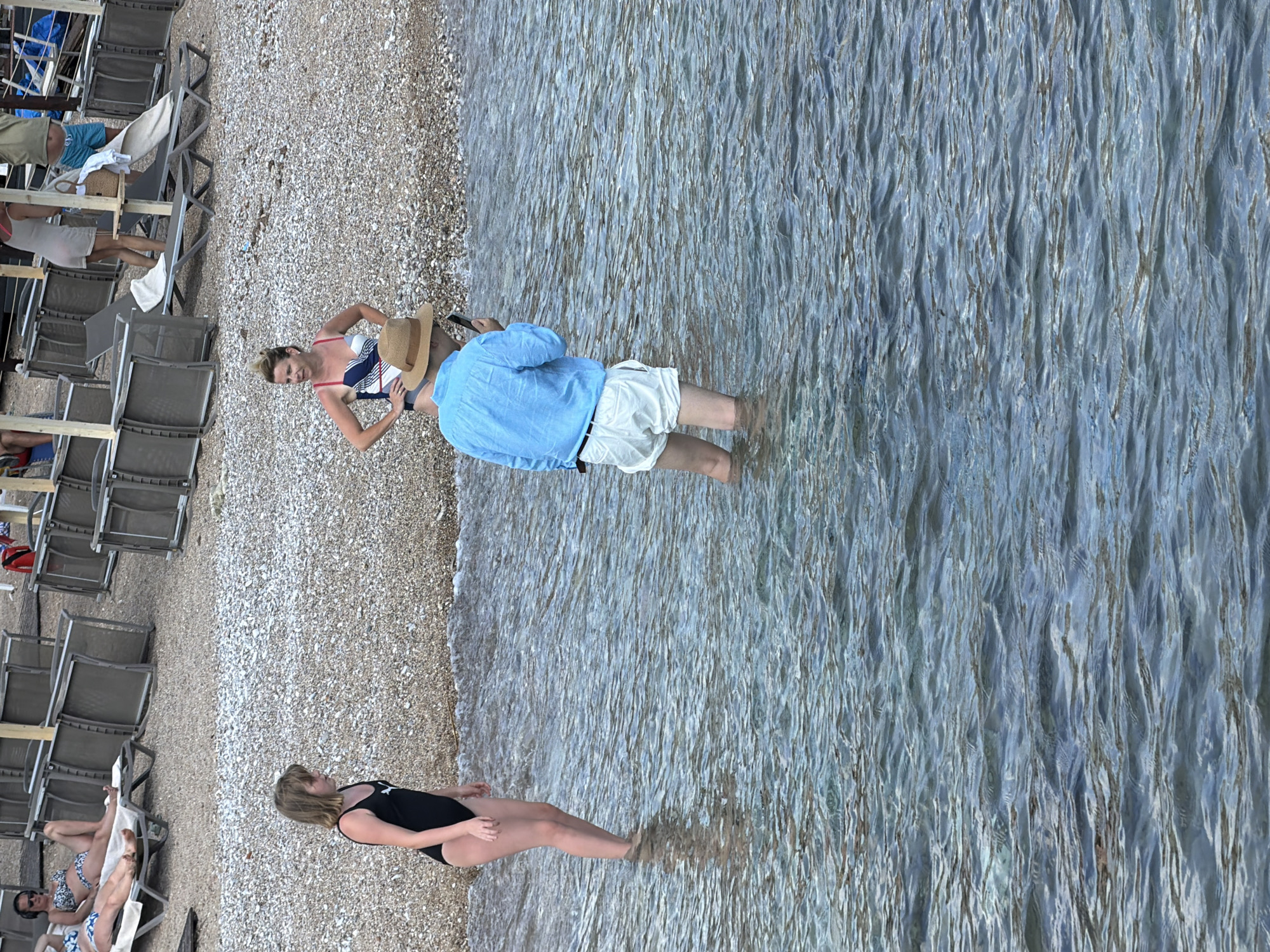 The image depicts a seaside scene on a pebble beach with loungers set up. In the foreground, a woman in a black swimsuit is standing in the shallow, clear water, with her legs partially submerged. Behind her, a man wearing a blue shirt and white shorts is also standing in the water, holding a phone and a straw hat. Further back, on the beach, a woman in a blue and white striped bikini is sitting on a lounger, holding a straw hat. She appears to be looking towards the water. The water is calm with a rippled surface, reflecting the light. The loungers are stacked and arranged neatly on the pebble beach. The scene suggests a relaxed atmosphere, likely during a warm, sunny day, as implied by the beach setting and people in swimwear. The location is identified as Budva, Montenegro. There is no visible text in the image.
