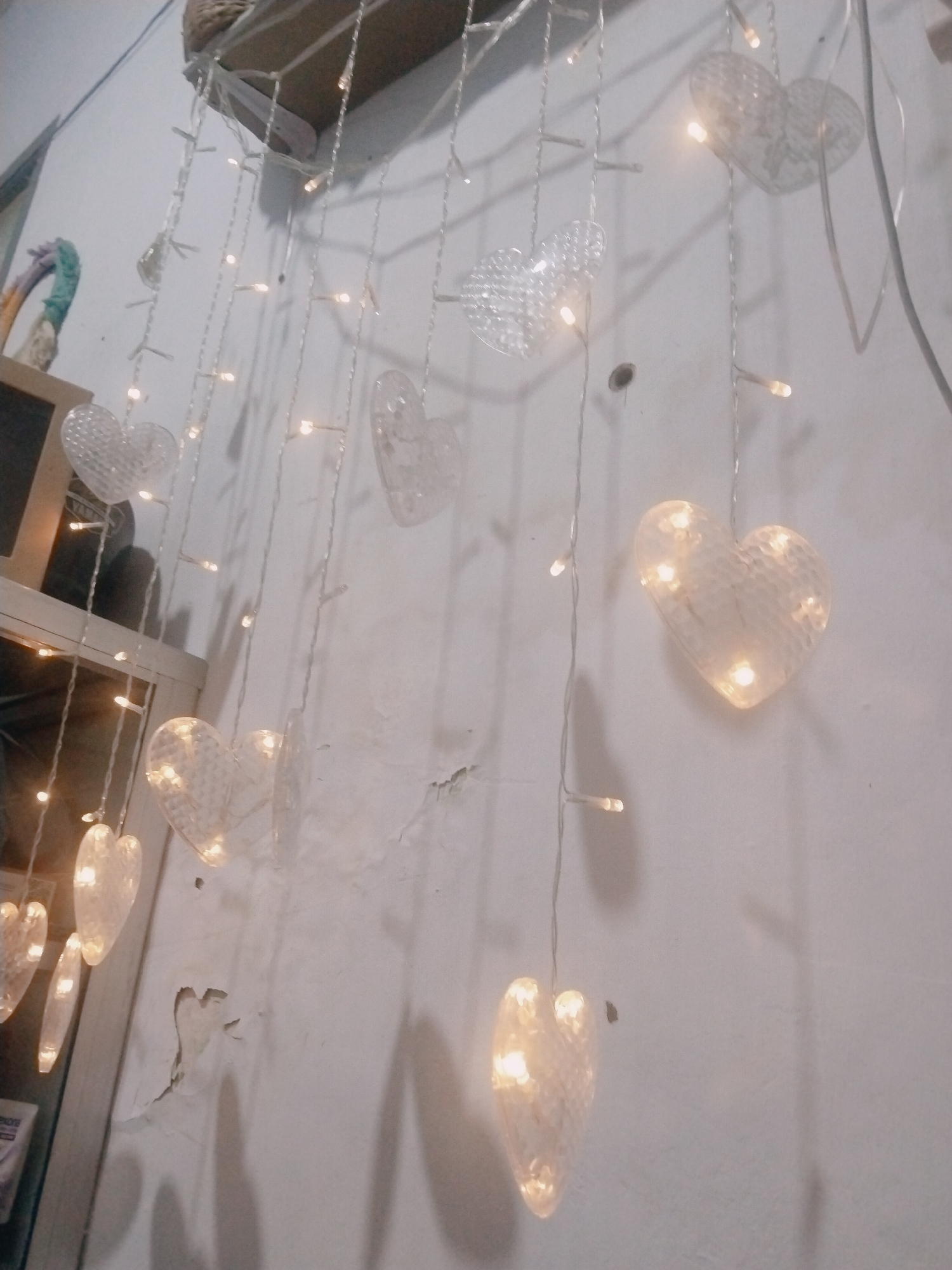 An indoor, upward-angled, medium shot captures a string of heart-shaped decorative lights hanging against a textured, white wall. The lights are warm white and illuminated, casting a soft glow. Several clear, textured heart-shaped ornaments are attached to the string, interspersed with the lights. In the upper left portion of the frame, a woven item, possibly a basket or shelf, is visible, along with a colorful, abstract sculpture. To the left, a shelf unit with a wooden frame contains some items, including what appears to be a box with text. The wall exhibits areas of chipped paint and texture. The scene is lit by the decorative lights and ambient light, creating subtle shadows on the wall. The location is not discernible from the image.