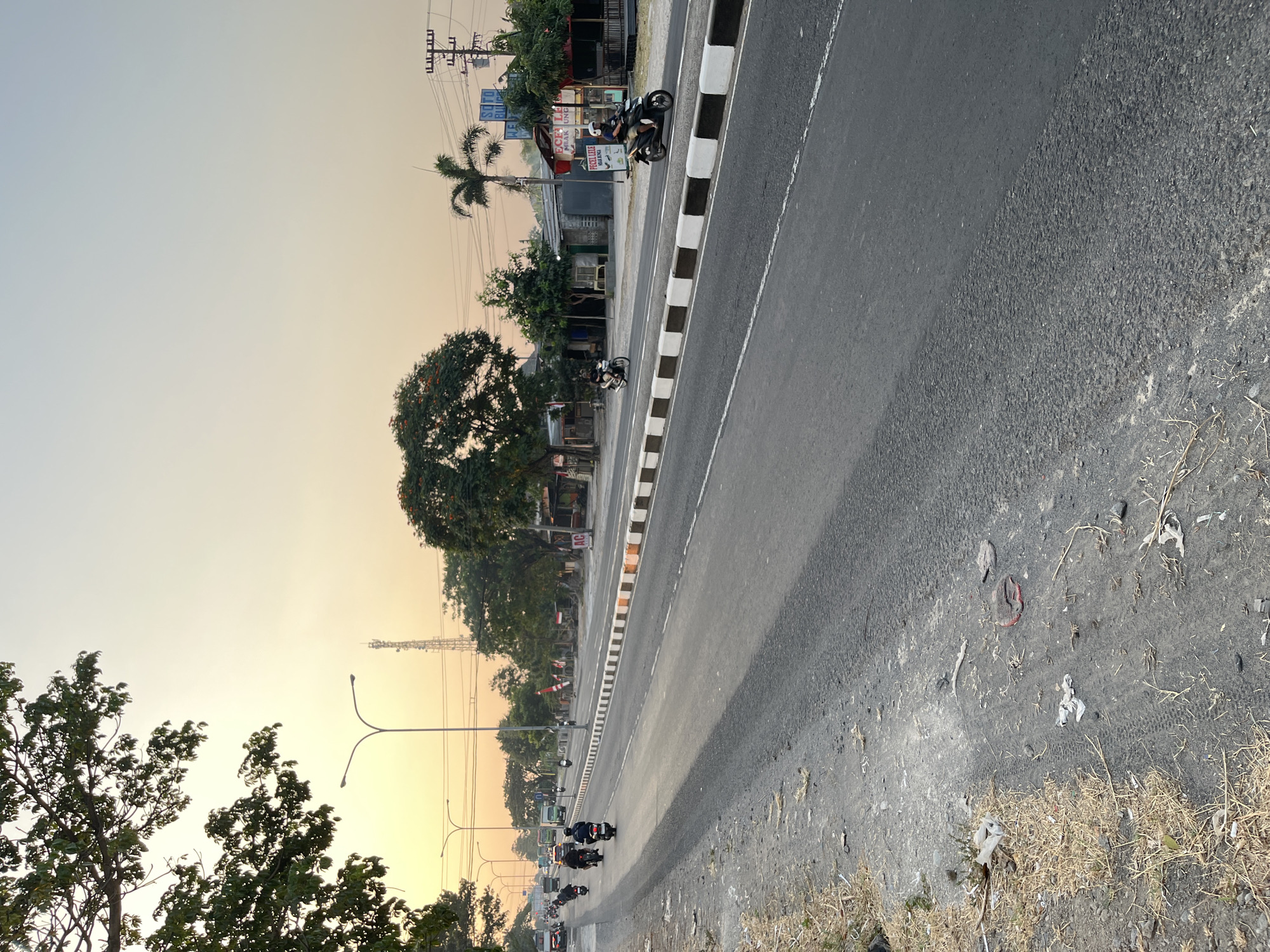 The image depicts a two-lane road in Surakarta, Indonesia, likely during the late afternoon or early evening due to the warm, golden light. On the left side of the road, a dirt shoulder is visible, littered with dry grass, debris, and some plastic waste. A large tree with green foliage arches over the left side of the frame. The road itself is paved, with a distinct white and black striped curb separating the traffic lanes from the shoulder.
Several motorbikes are in motion on the road, with riders dressed in casual clothing and helmets. Some are traveling in the same direction, while others appear to be passing. In the background, on the right side of the road, there are several small buildings, some appearing to be shops or food stalls. Signs are visible on these buildings, with text in Indonesian, including "SOTO," "Pecel Lele," and "Mbak Ungu." Electrical poles and wires are also present along the roadside.
Further down the road, more traffic is visible, including trucks and cars. A tall communication tower stands in the distance against the hazy, light orange sky. The overall impression is of a busy, functional road on the outskirts of a city, characterized by local commerce and everyday traffic.
