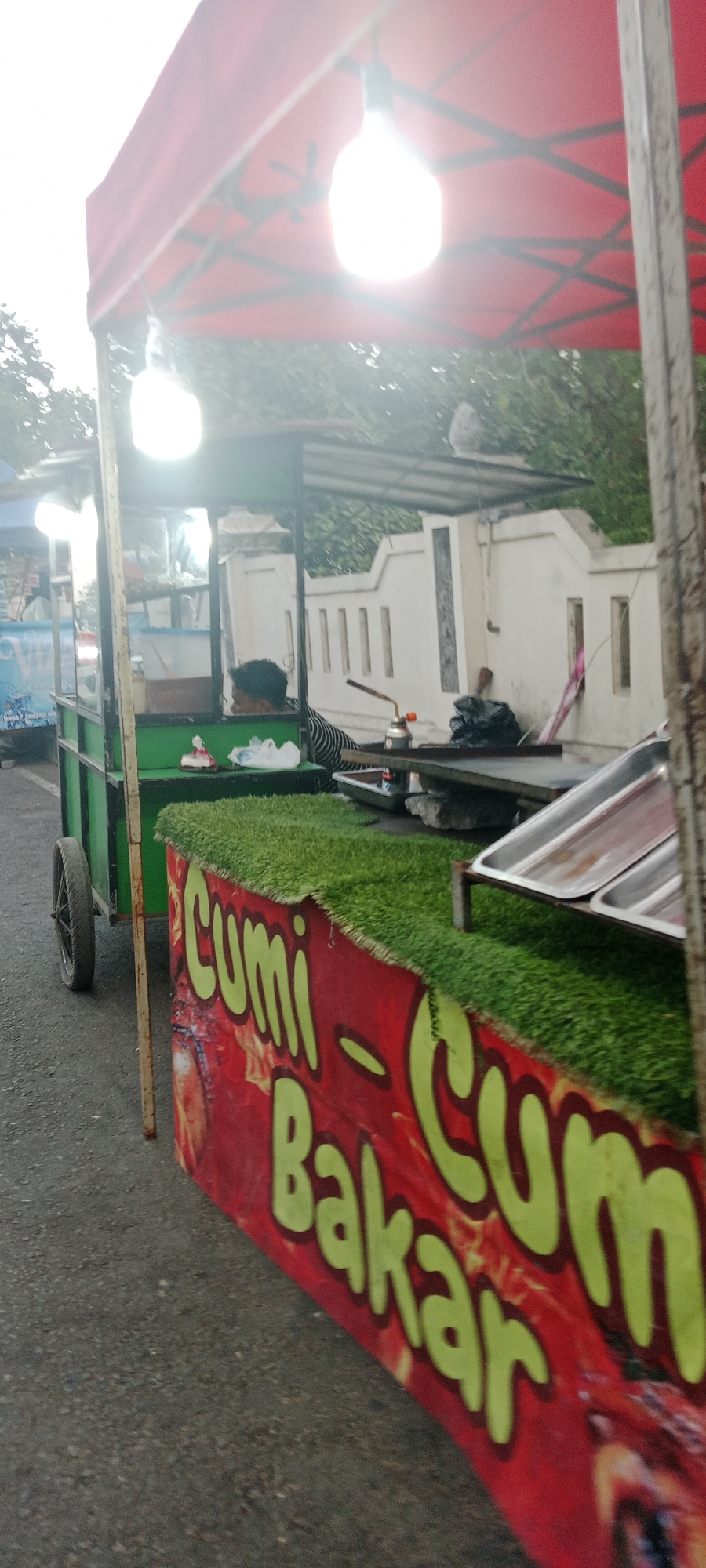 A street food cart is parked on the side of a road. The cart has a red canopy and is adorned with green artificial grass along the top edge of the front panel. The front panel displays a red banner with large, yellow text that reads "Cumi - Cumi Bakar," which translates to "Grilled Squid." The cart appears to be preparing food, with a person in a striped shirt visible inside, and a blowtorch resting on a metal surface. Several stainless steel trays are visible on the cart, suggesting food preparation or serving. The background shows a white, textured wall with vertical slats and some greenery, indicating an outdoor urban setting. The overall scene suggests a food stall operating in Ngemplak, Indonesia.