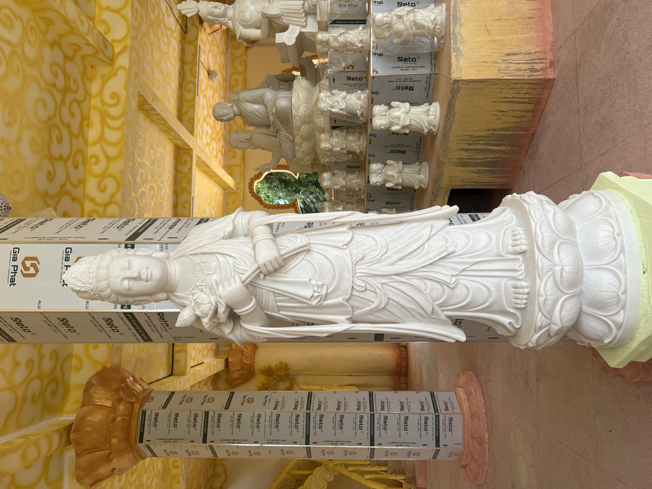 An indoor religious facility in Đăk Mar, Vietnam, is depicted, dominated by a tall, white marble statue of a female deity, likely Guanyin, standing centrally and facing forward. The figure, adorned with an elaborate headdress and flowing robes, holds a lotus flower in her left hand and stems in her right, resting on a multi-tiered lotus pedestal. The ceiling and upper architectural elements are painted yellow and gold with swirling cloud patterns. Support pillars in the foreground and mid-ground are covered with white aluminum composite panels featuring "Gia Phat Alu" and "Reto" branding. To the right of the central statue, a raised platform with a discolored yellow-and-brown finish supports multiple white statues. These consist of larger seated Buddhist figures on lotus bases and multiple smaller statues, some enclosed in clear plastic, depicting praying or gesturing figures arranged in rows. An arched opening in the distant background provides a view of green foliage. The floor consists of light-colored, patterned tiles. To the far left, a golden pillar with a lotus-shaped capital and yellow decorative elements are present.