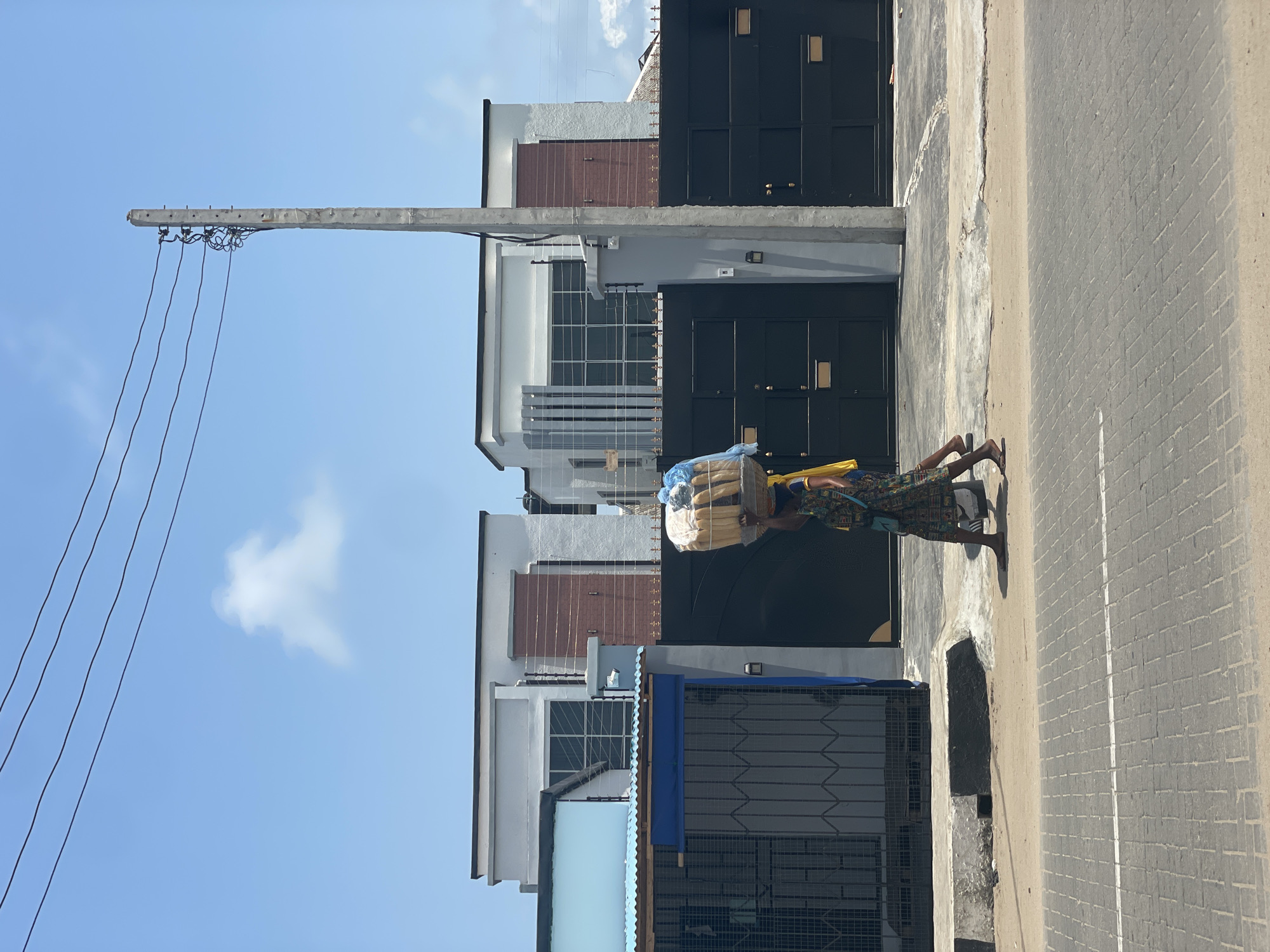 This daytime image depicts a street scene in Badagry, Nigeria. A person is seen walking on the right, carrying a large stack of light-colored, rectangular packages, possibly bread or provisions, topped with a blue plastic wrap, balanced on their head. They are dressed in a colorful, patterned outfit and wear sandals.
To the left, a series of modern buildings line the street, characterized by dark gates, white and grey walls, and some featuring brown accents, windows with grilles, and balconies. A tall utility pole with numerous overhead wires dominates the left side of the frame, extending into the clear blue sky, which has a few scattered white clouds, indicating sunny weather. The road surface appears to be paved with a textured pattern, resembling interlocking bricks, and a narrow sidewalk or dirt path runs alongside the buildings. No visible text is legible in the image.