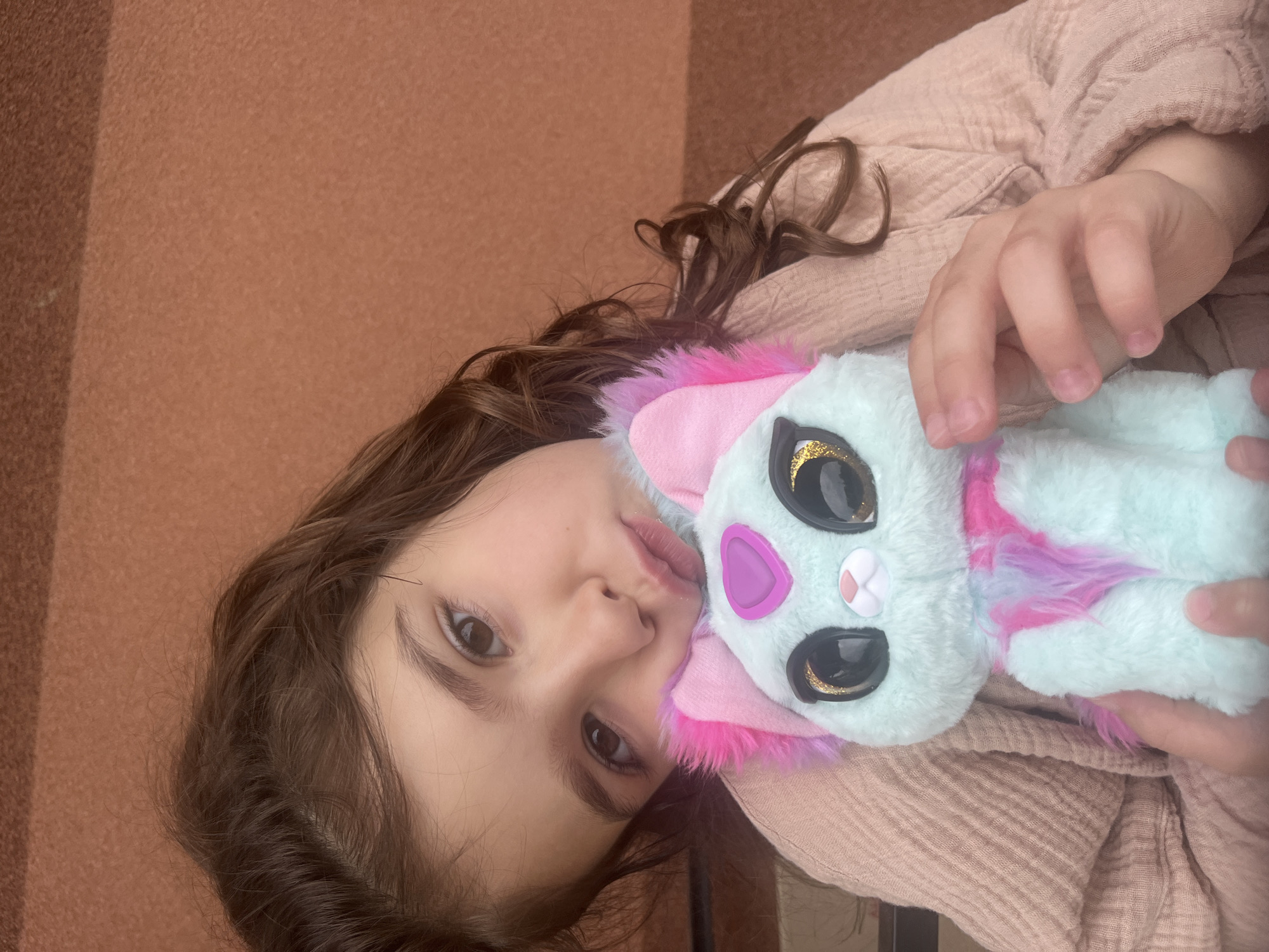 A young child, with dark brown, wavy hair and light brown eyes, is positioned in a medium close-up in Onokivtsi, Ukraine, gazing directly at the camera with a puckered lip expression. The child's head is tilted left, resting against a plush toy held with both hands. The child is wearing a light peach-colored, textured, long-sleeved garment. The plush toy, a stylized cat-like figure, is predominantly mint green, featuring large black eyes with gold glitter accents, pink and purple furry ears, a pink heart emblem on its forehead, and additional pink and purple fur accents on its chest. The background consists of an unadorned, textured, reddish-brown surface, possibly a wall, exhibiting a faint horizontal line across its upper third. The scene is evenly lit.