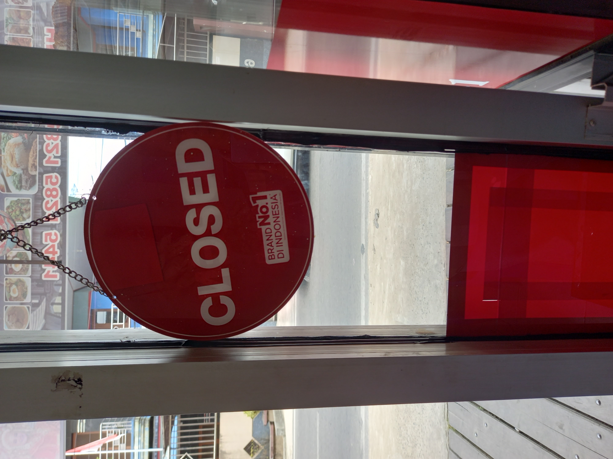 A red circular "CLOSED" sign hangs by a chain in the doorway of a business, partially obscuring a view of the street and buildings outside. The sign displays white lettering that reads "CLOSED" prominently, with "BRAND No.1 DI INDONESIA" below it. The doorway appears to be made of glass and metal, with wooden elements on the left side and red reflective surfaces on the right. Through the glass, a street with vehicles and buildings can be seen, suggesting an urban setting. The overall impression is that of a closed establishment in Samarinda, Indonesia.