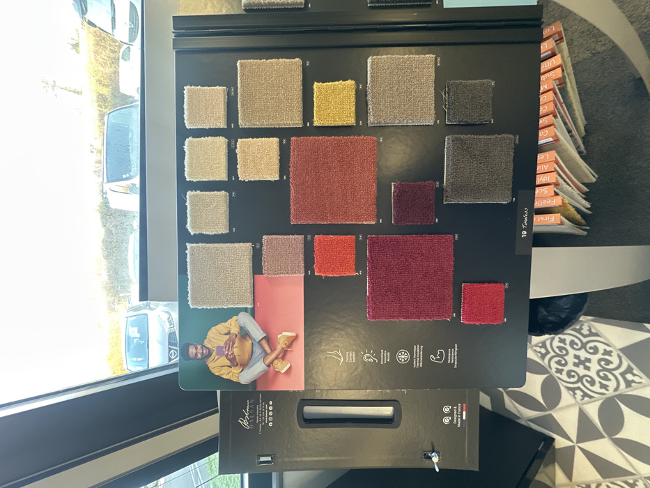 The scene is an interior view of a commercial space in Sainte-Geneviève-des-Bois, France. A large, black, upright display board occupies the central and right portions of the frame, showcasing numerous square and rectangular carpet swatches. These swatches are presented in a variety of colors, including beige, cream, pink, orange, red, burgundy, yellow, light grey, and dark grey, each with a numerical identifier. The upper left quadrant of this board features a printed image of an adult male with a beard, wearing a yellow hooded sweatshirt and grey trousers, seated cross-legged on a red background while holding a smartphone. Below this image, icons illustrate product features with accompanying French text, such as 'Confort' and 'Résistance'. The bottom center of the board displays the text '19 Timeless'. To the immediate left, a narrower black display panel is partially visible, indicating 'BALSAN' branding and 'Designed & Made in France'.
Through a large window in the background, an outdoor parking area is visible under bright daylight. Several light-colored vehicles, including vans and cars, are present, with green foliage visible beyond them. The interior floor exhibits a geometric pattern composed of grey, white, and black tiles. Partially visible beneath the main display board are a stack of brochures with colorful, labeled tabs (e.g., 'First', 'Feeling', 'Idylle') and a dark object on the floor.