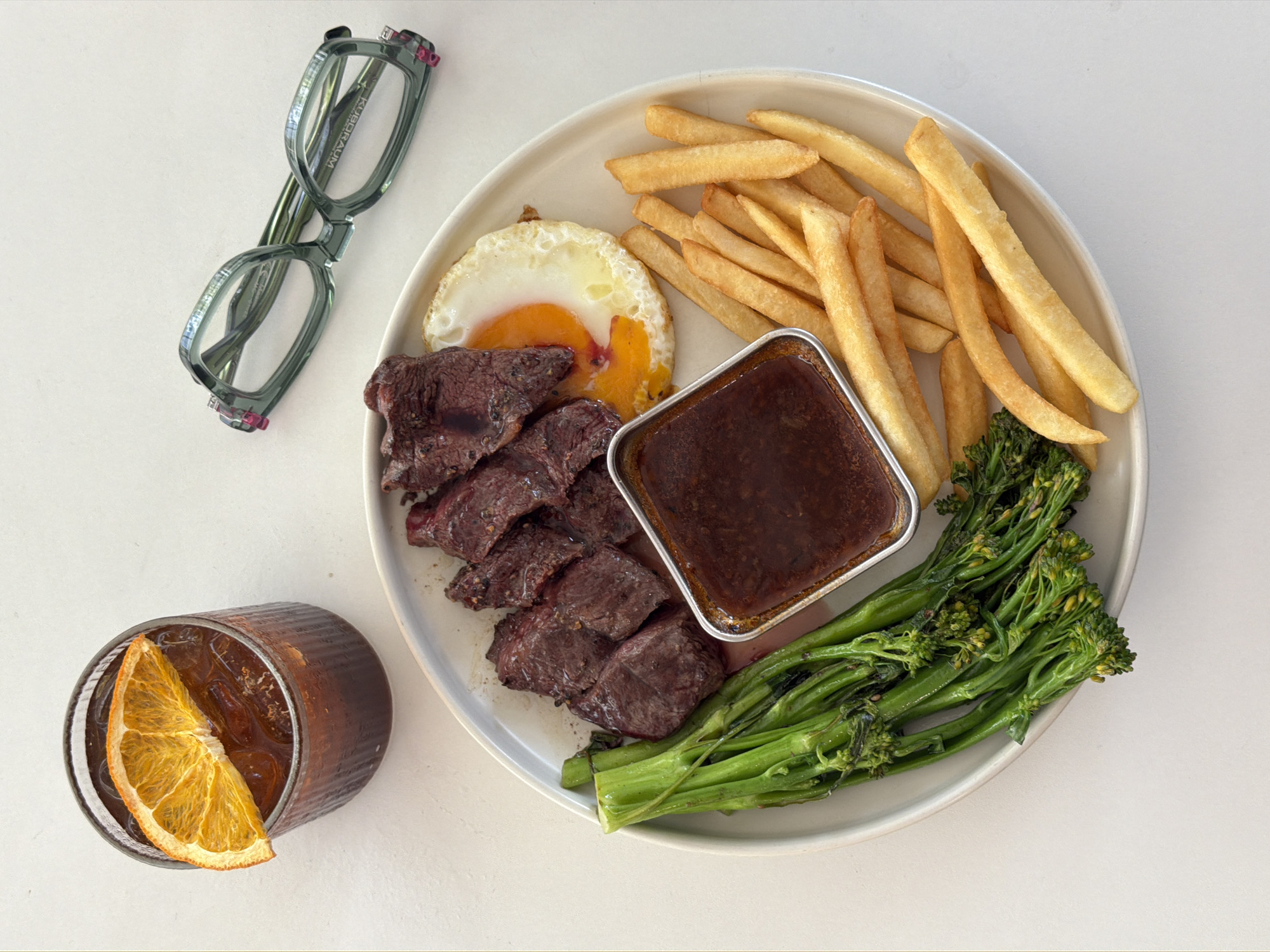 A high-angle shot depicts a plate with a meal consisting of steak, french fries, broccolini, a fried egg, and a square dish of gravy. To the left of the plate are a glass with an orange slice and a pair of glasses. The image appears to be taken indoors, on a white surface. The specific location in Đà Nẵng, Vietnam is not identifiable from the image.