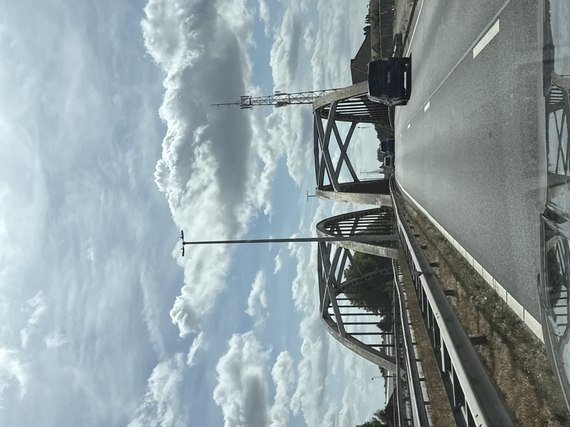 The image shows a view from a vehicle driving on a highway towards a bridge. The bridge has a distinctive arched design with metal supports. There are several cars and a van on the highway. On the right side of the highway, there is a tall telecommunications tower. The sky is partly cloudy. The environment appears to be a suburban or rural setting due to the presence of dry grass and vegetation on the sides of the road. There are no visible people. The scene is captured on a bright, overcast day.
