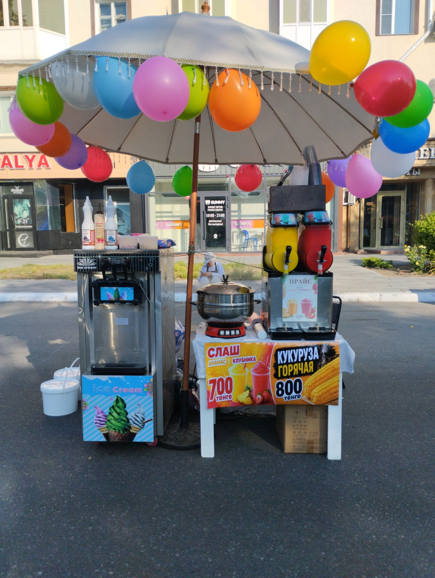 An outdoor scene depicts a portable food and beverage stand on an asphalt surface, likely in the city of Kostanay, Kazakhstan, given the signage. The stand is positioned in front of a building with storefronts, one of which displays the name "ANTALYA". A large, gray umbrella, adorned with colorful balloons, is erected above the stand. The stand itself features an ice cream machine on the left, with a sign for "ice Cream" and an image of ice cream cones. To the right of the ice cream machine are two dispensers filled with yellow and red beverages, and a steamer pot on a stand with a sign displaying prices in Kazakhstani Tenge. A cardboard box with Chinese writing is visible beneath the right side of the stand. The environment suggests a commercial street setting, with a person visible in the background sitting on the ground.
