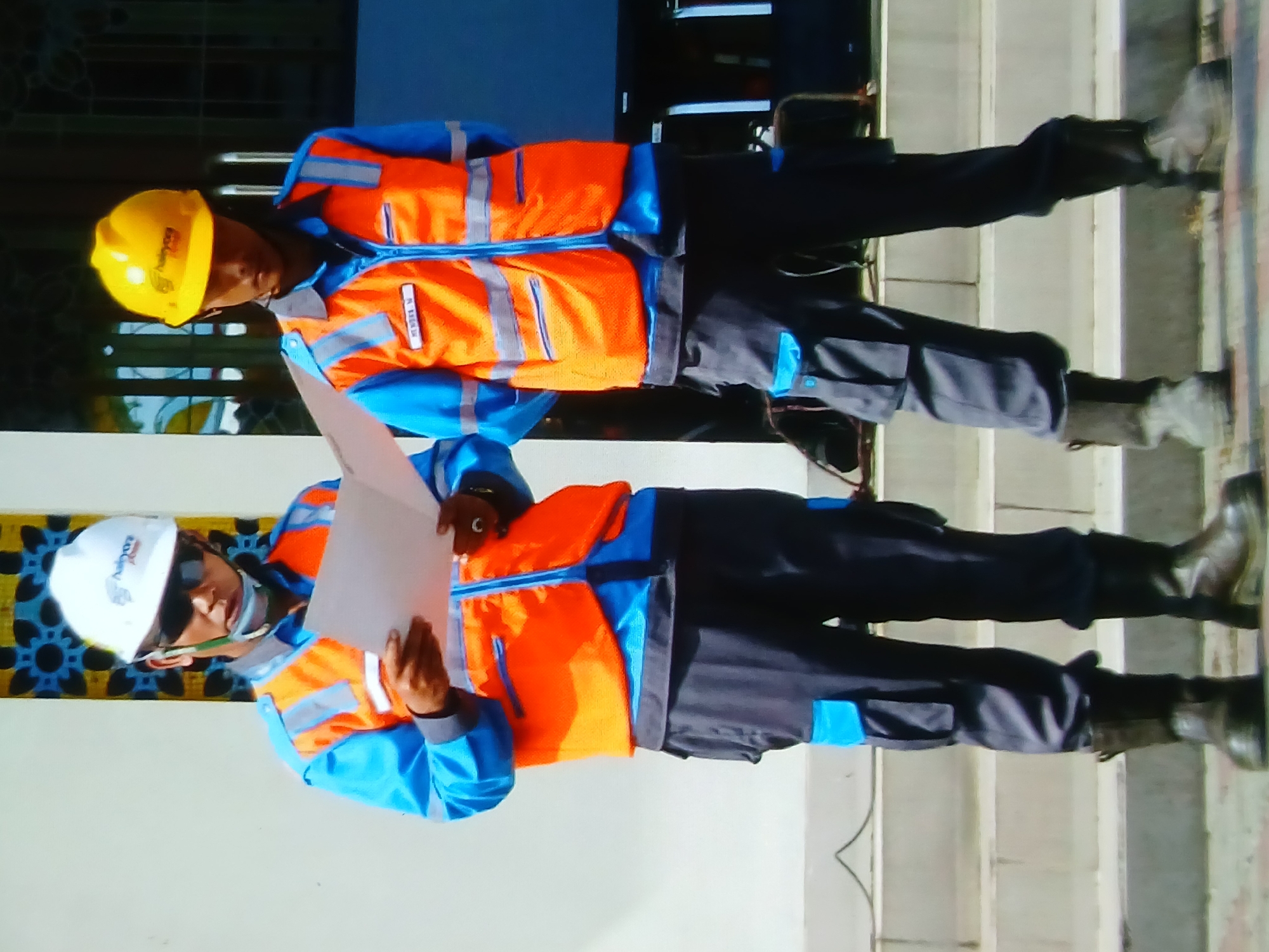 Two individuals, dressed in work uniforms consisting of blue shirts, orange safety vests with reflective strips, and dark grey work pants with blue accents, stand on a set of concrete stairs. Both individuals are wearing hard hats, one white and the other yellow. The individual on the left wears sunglasses and holds a white paper document. The individual on the right has a name tag on their vest that reads "MEIDEN M". The background features a white wall with decorative elements and a dark structure with vertical lines. This scene is located in Setia Bakti, Indonesia.