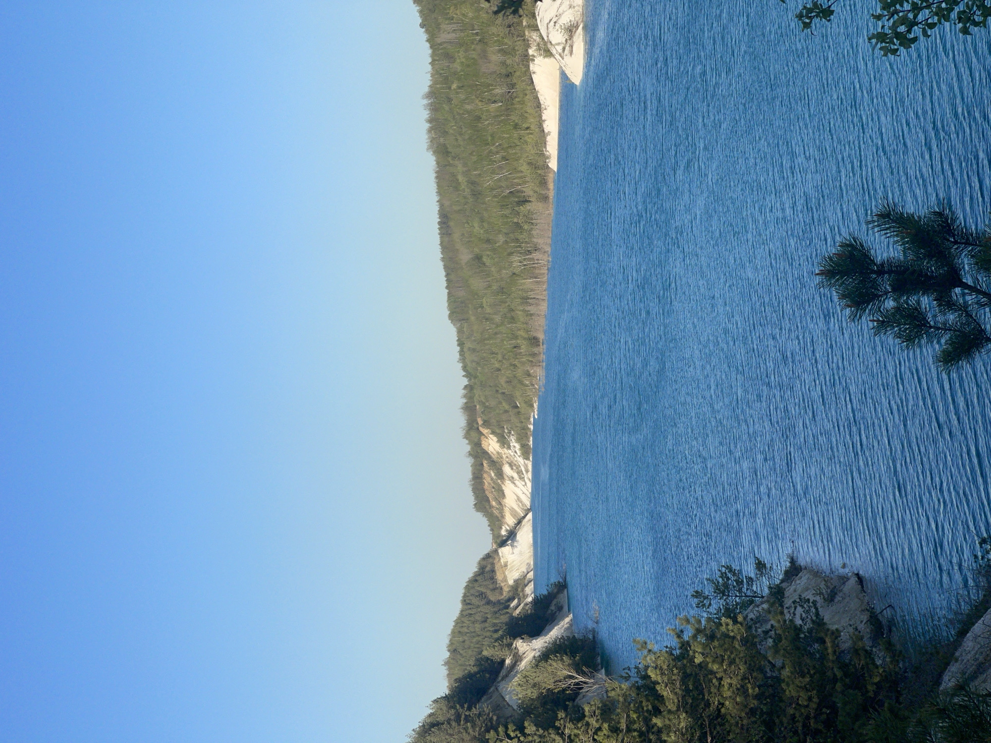 The image shows a body of water, likely a lake, with a wooded shoreline. The water is a bright blue color and appears to have slight ripples. The shoreline consists of green trees, and some exposed white cliffs or rocks. The sky is a clear, light blue. The exact location is Краснасельскі сельскі Савет, Belarus.